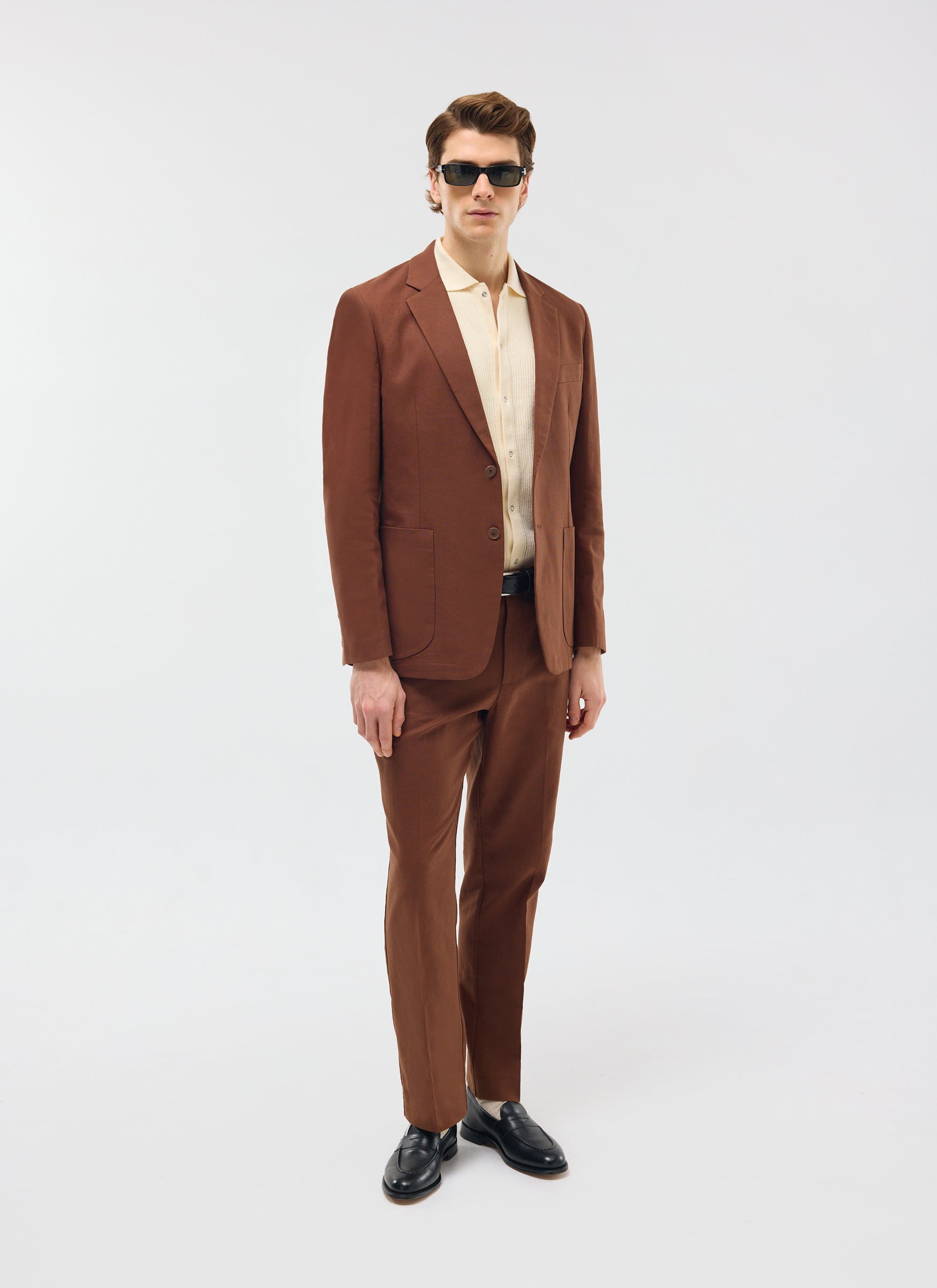 A model wears the Percival Tailored Linen Trousers in brown with the matching blazer, a cream knit shirt and sunglasses.