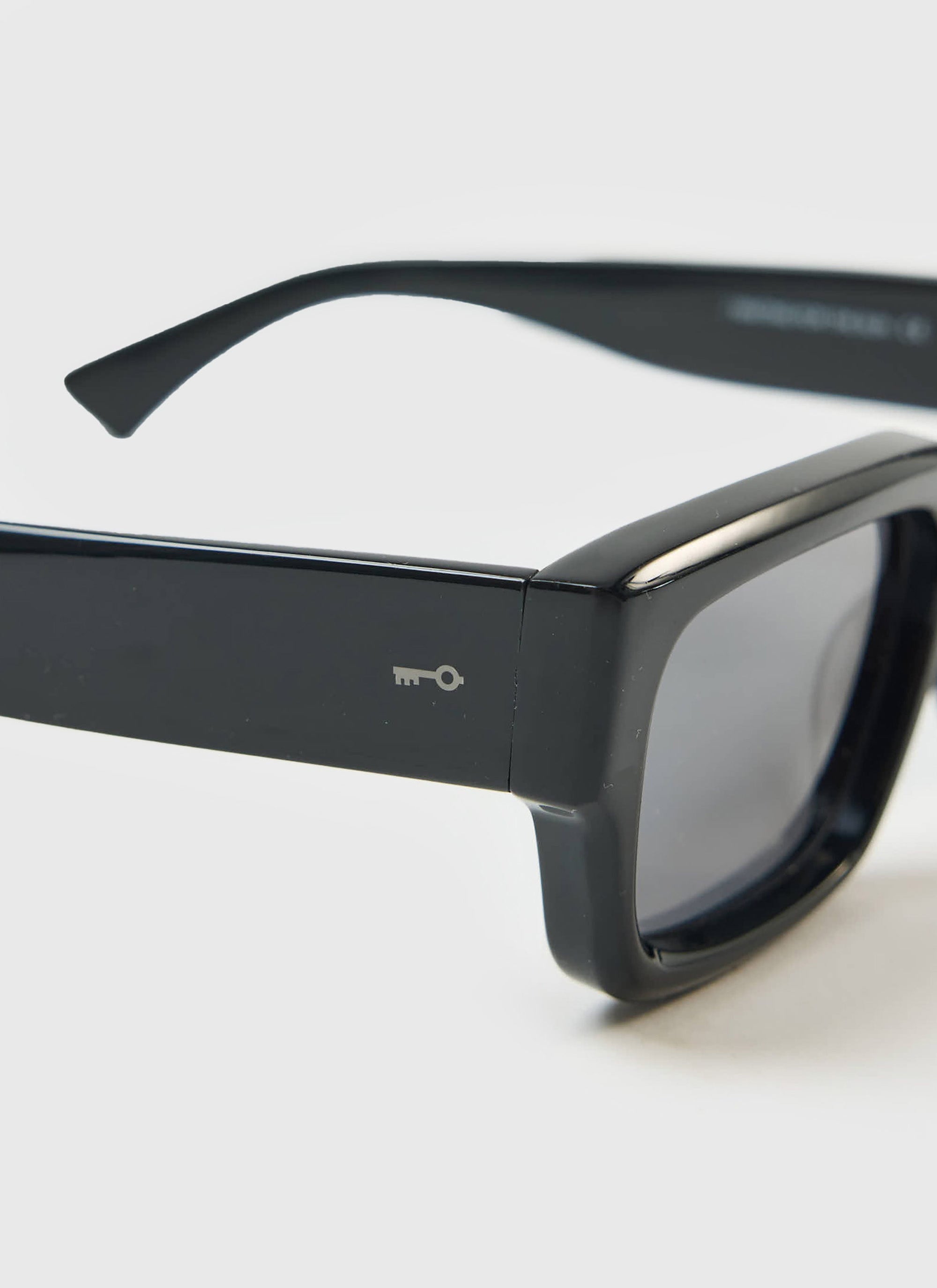 A close-up of the glossy black frame of the P002-E2 sunglasses by Percival, showing the subtle key logo branding.