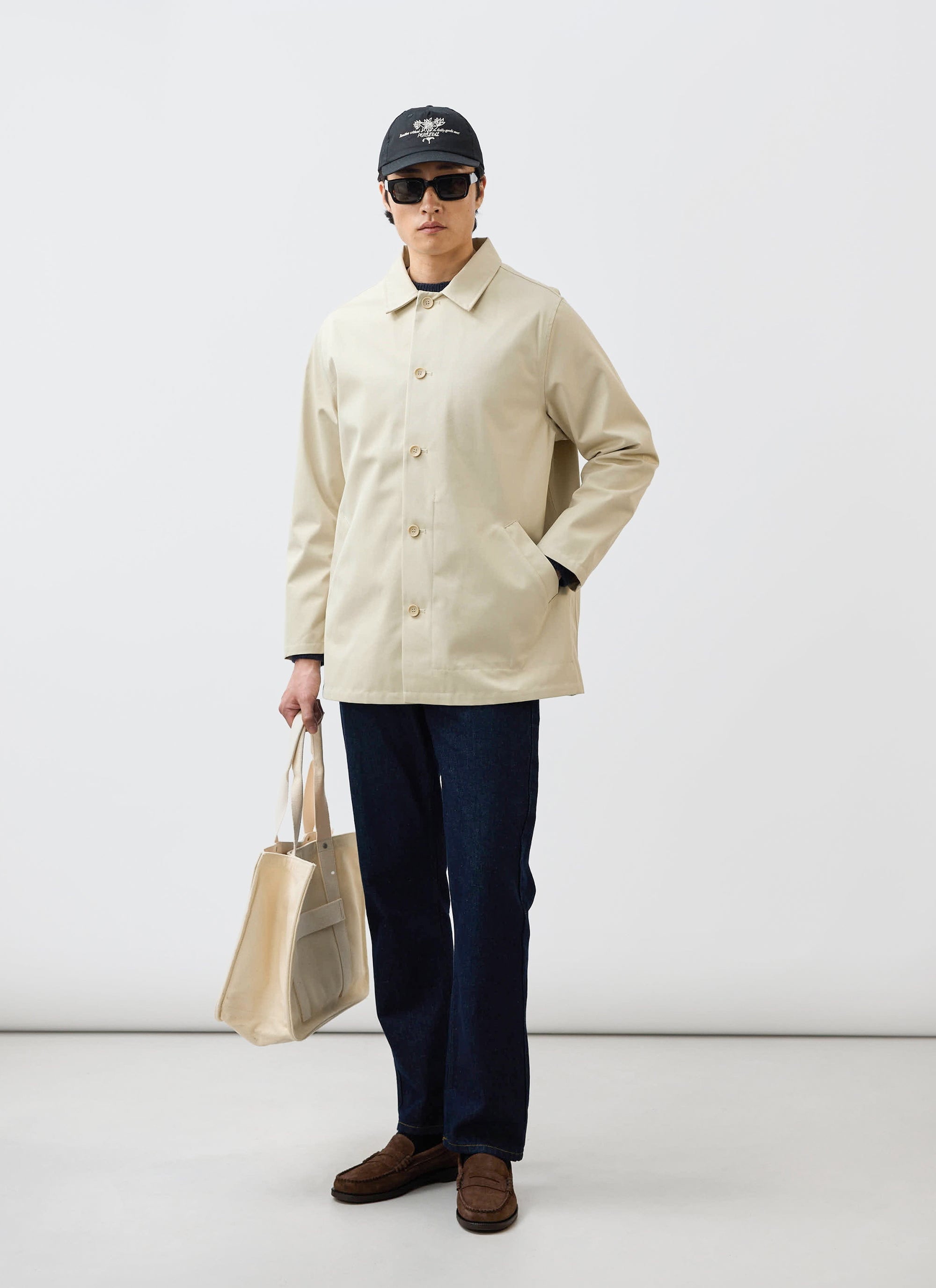 A man models the Percival Sherlock Short Trench Coat in stone, paired with dark jeans, sunglasses, and a tote bag.