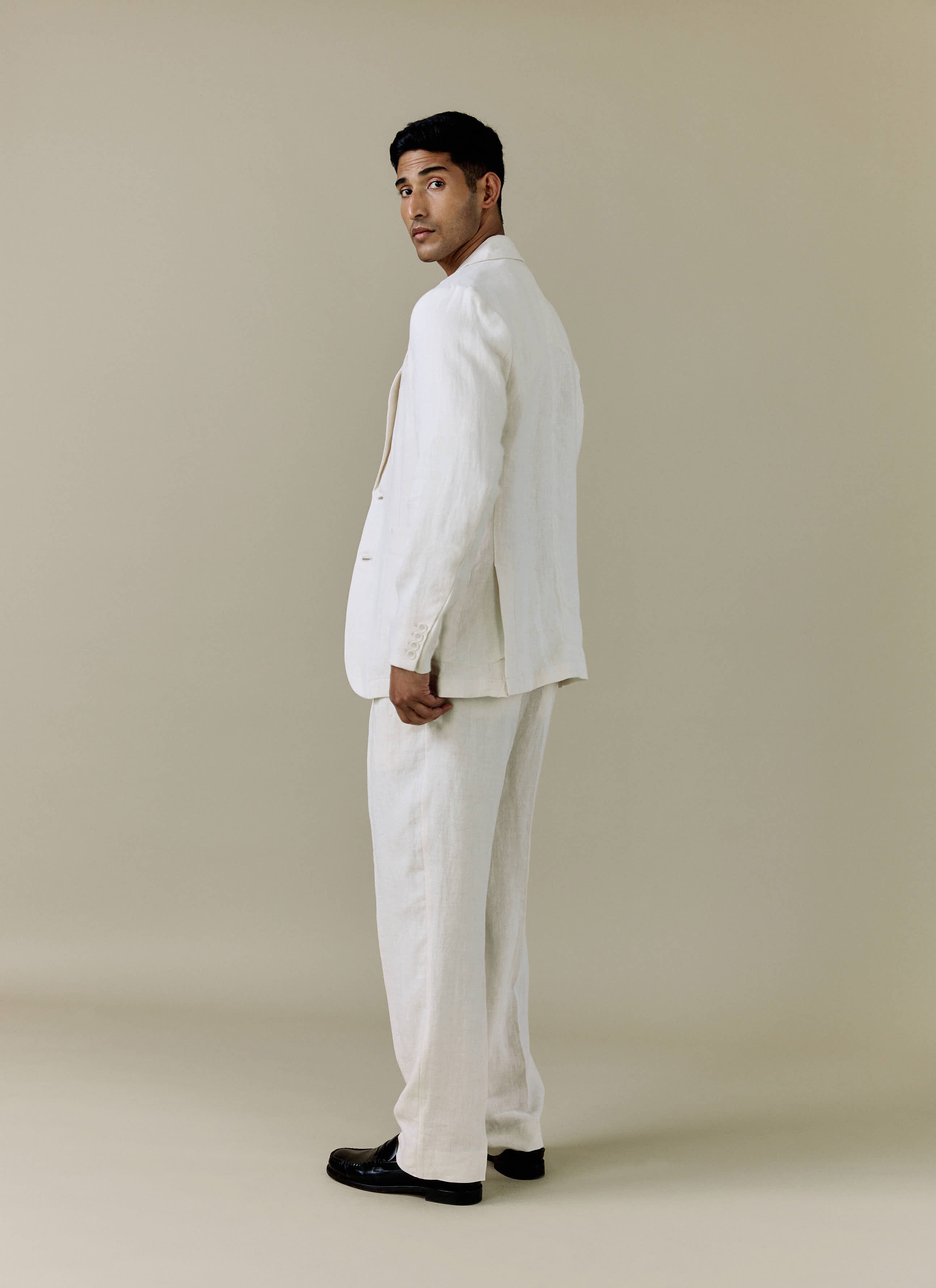 A model looks over his shoulder wearing the Percival Tailored Linen Blazer and matching trousers in cream.