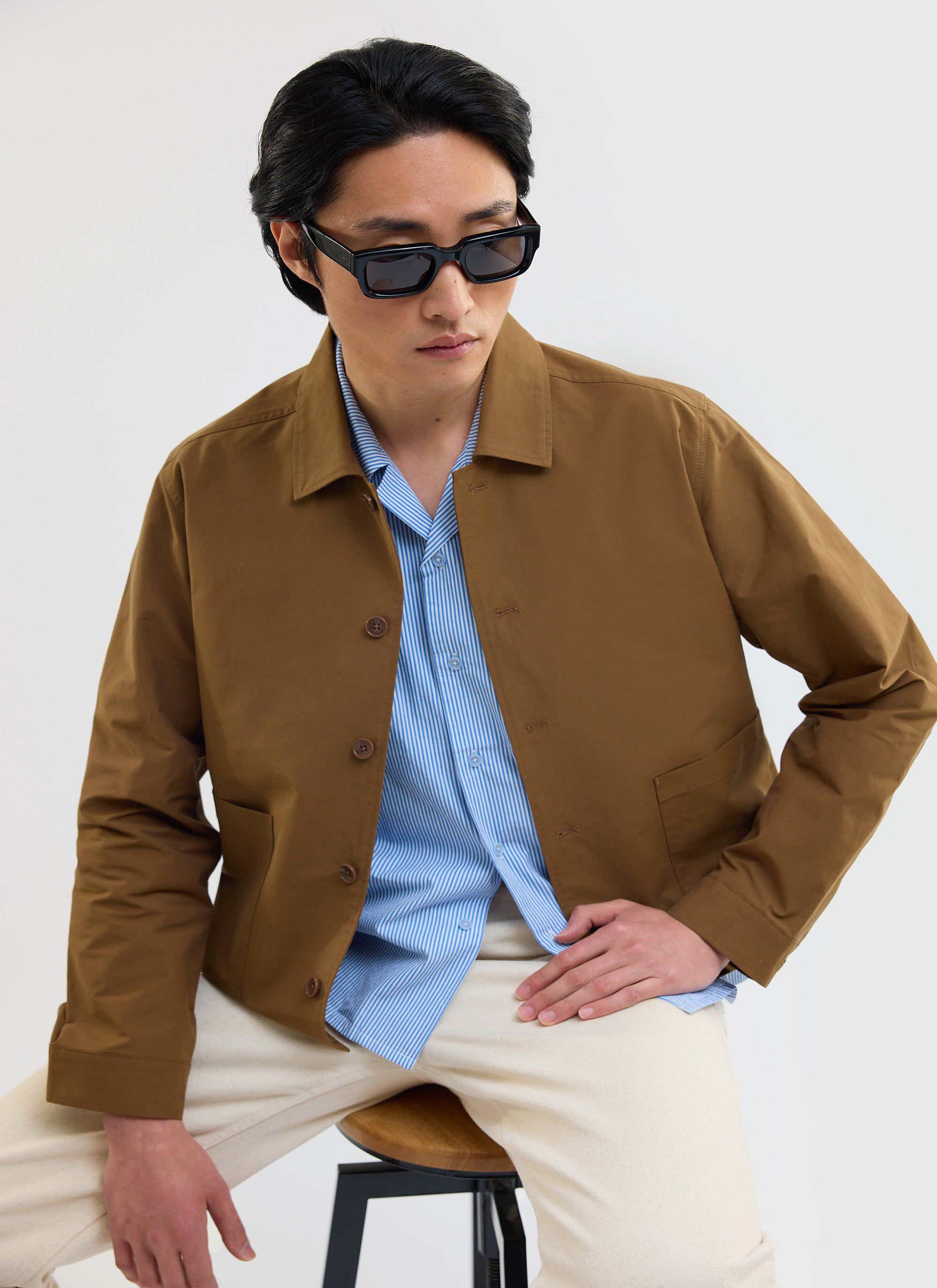 A model wears the Percival Boxy Cropped Pocket Overshirt in brown over a blue striped shirt and cream trousers.