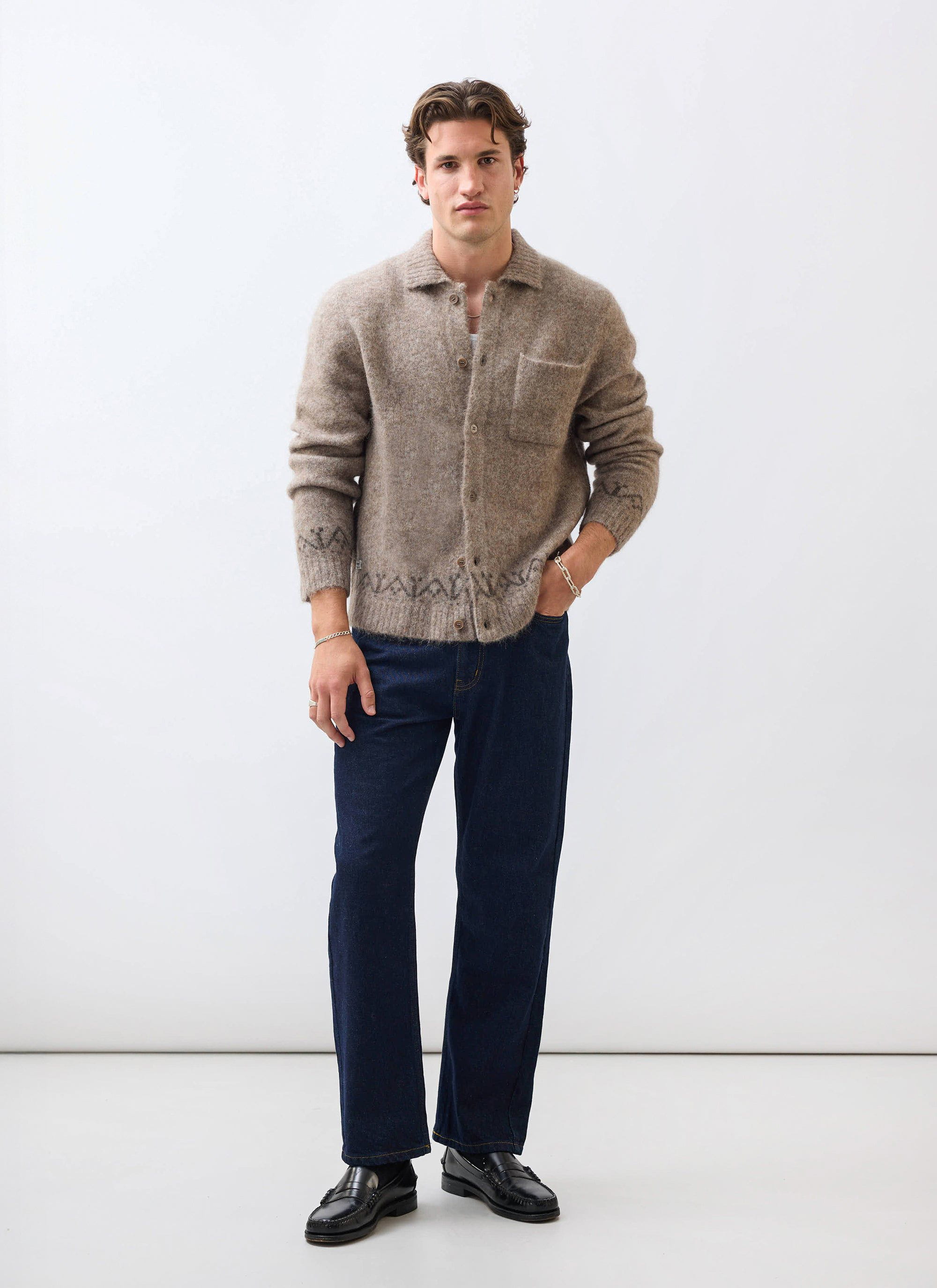A man models the Percival Novel Collared Cardigan in oak, paired with dark wash jeans and black loafers in a studio.