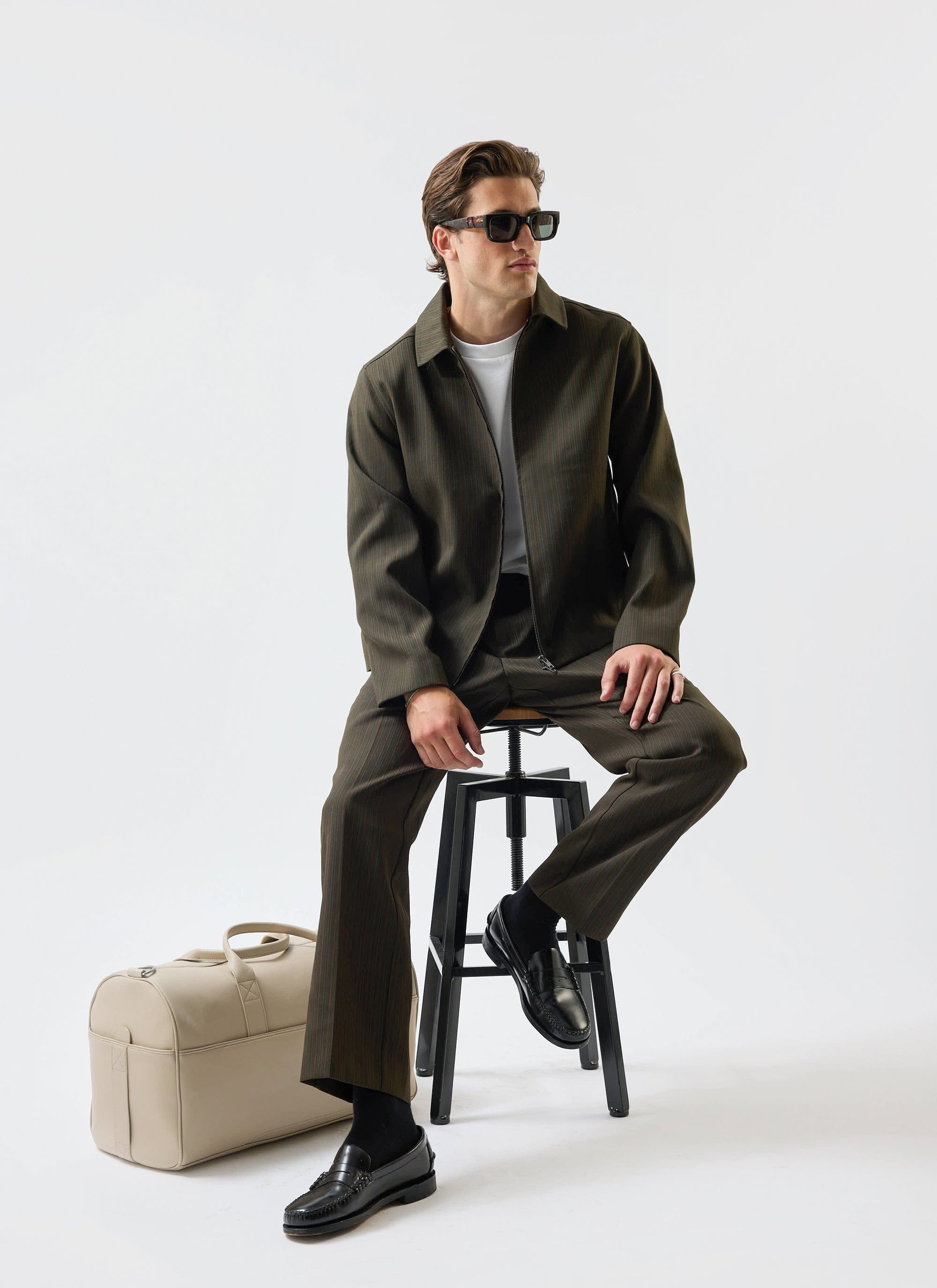 A model wears the Percival Chapter Zip Jacket in Forest Green with matching trousers, seated on a stool in a studio setting.