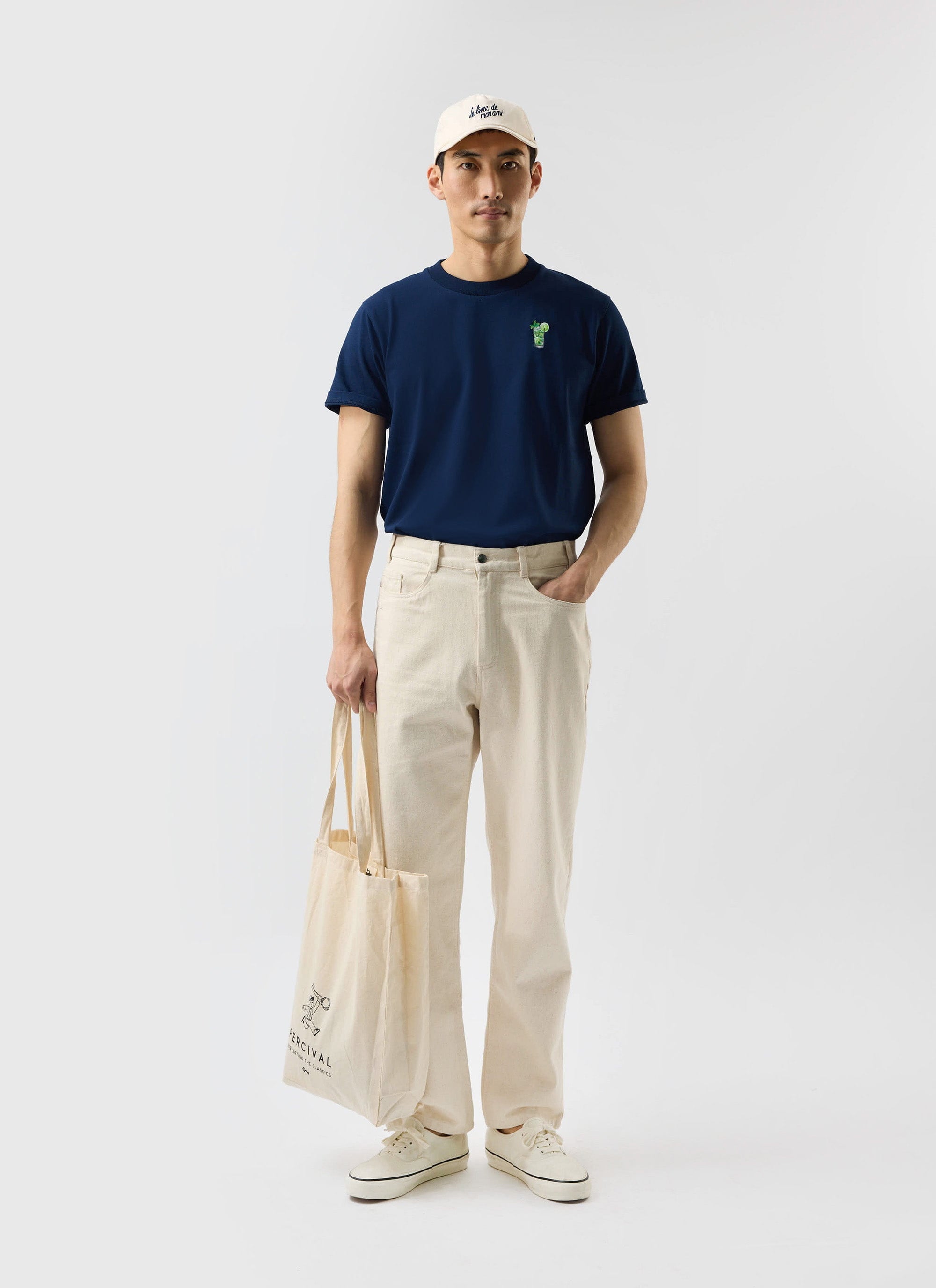 A model wears the navy organic cotton Mojito T-Shirt from Percival, styled with off-white trousers and a tote bag.
