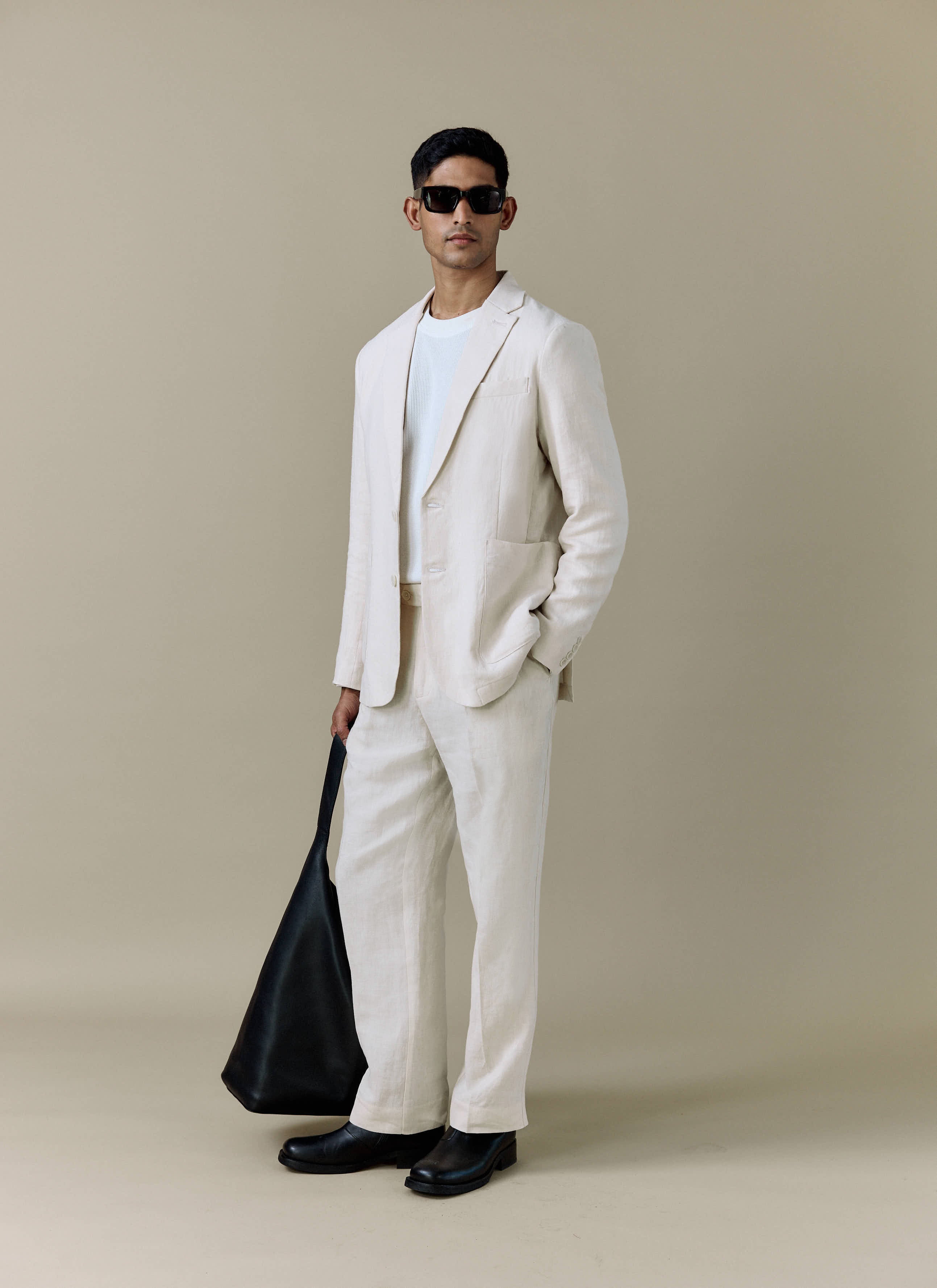 A model wears the cream linen suit from Percival, styled with a white t-shirt, sunglasses and a black bag.