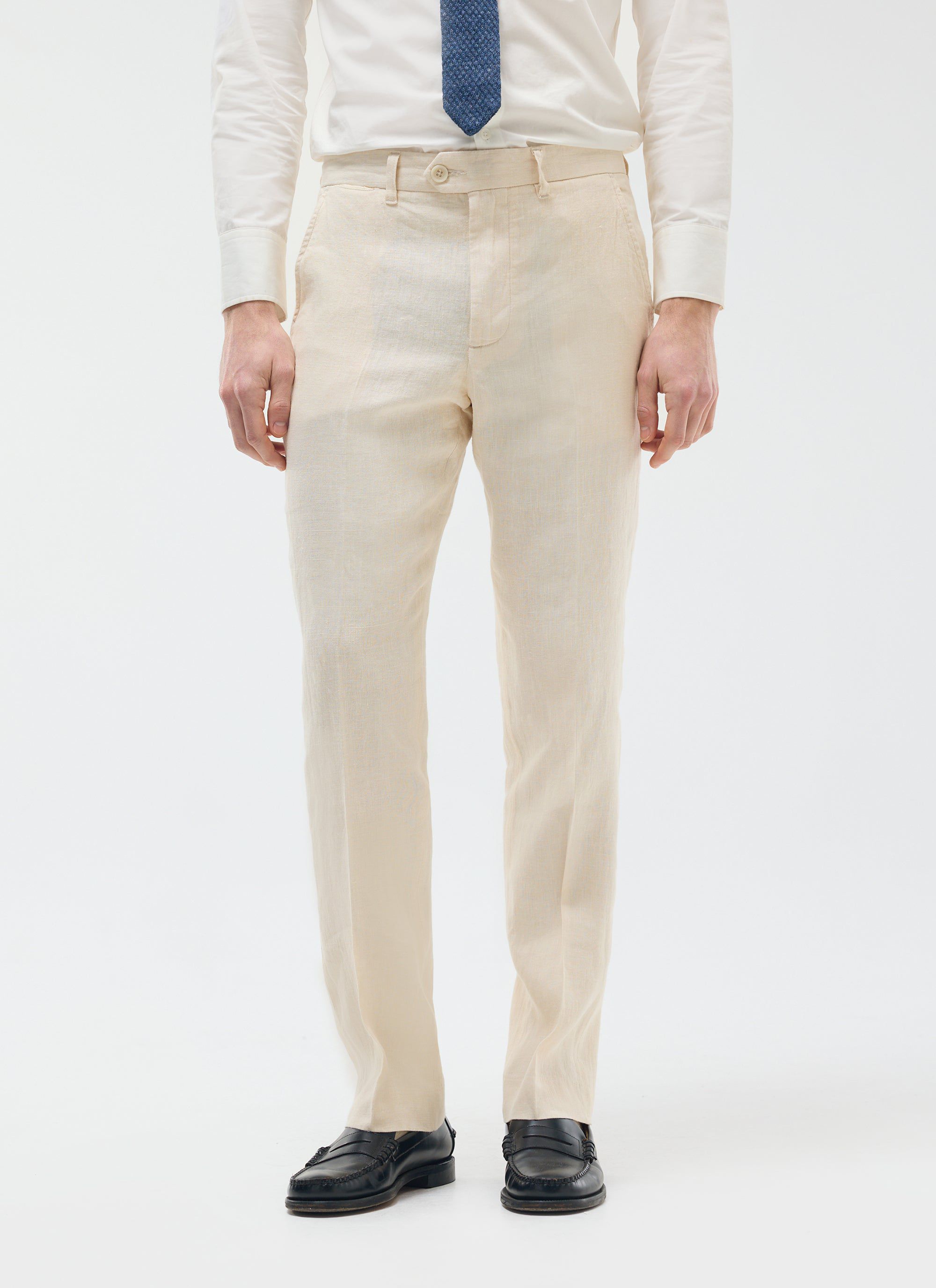 A model wearing Percival cream tailored linen trousers with a white shirt, blue tie and black loafers on a white background.