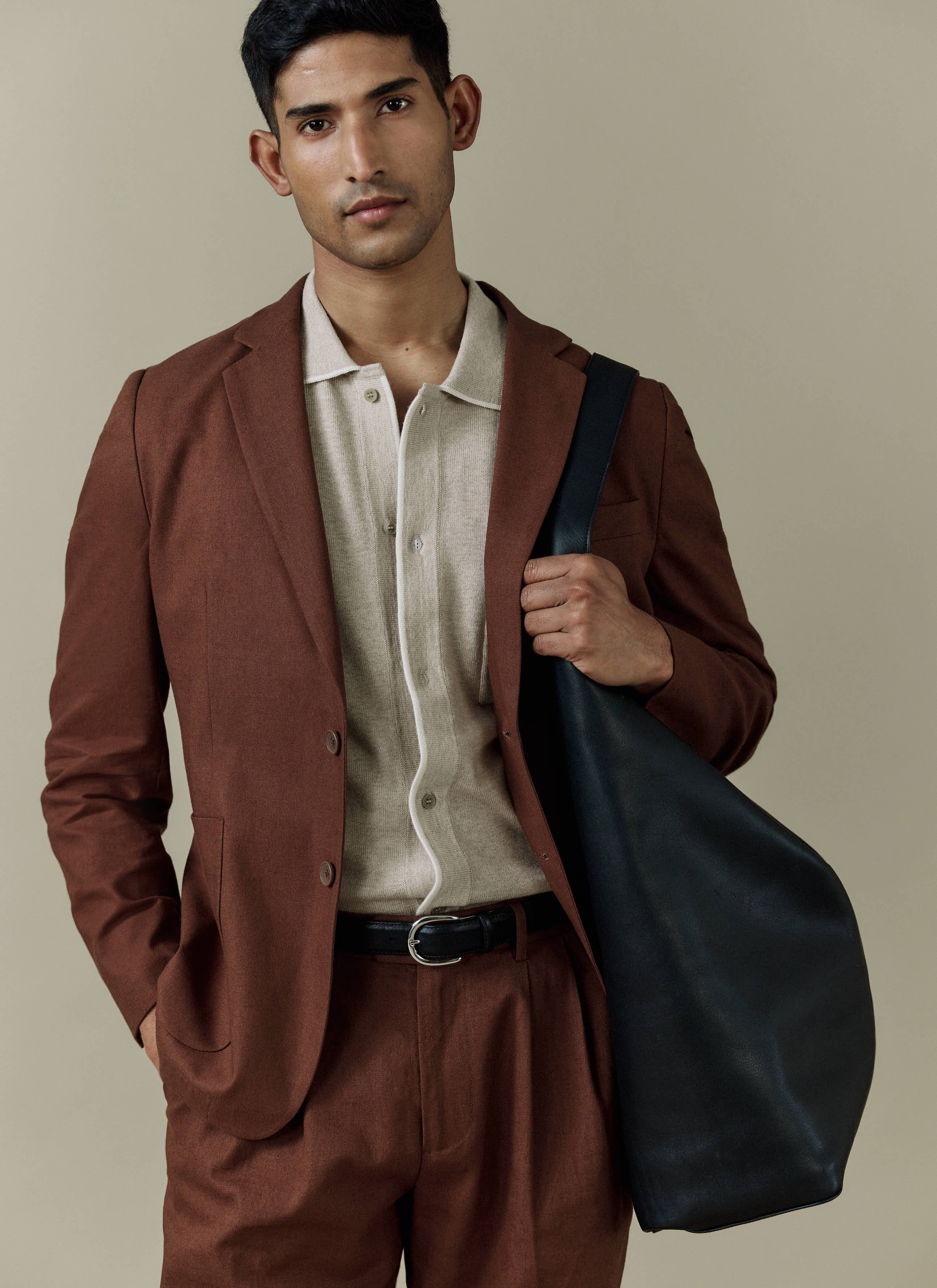 A model wears the Percival Tailored Linen Blazer in brown, styled with a cream knit shirt and a large black bag.
