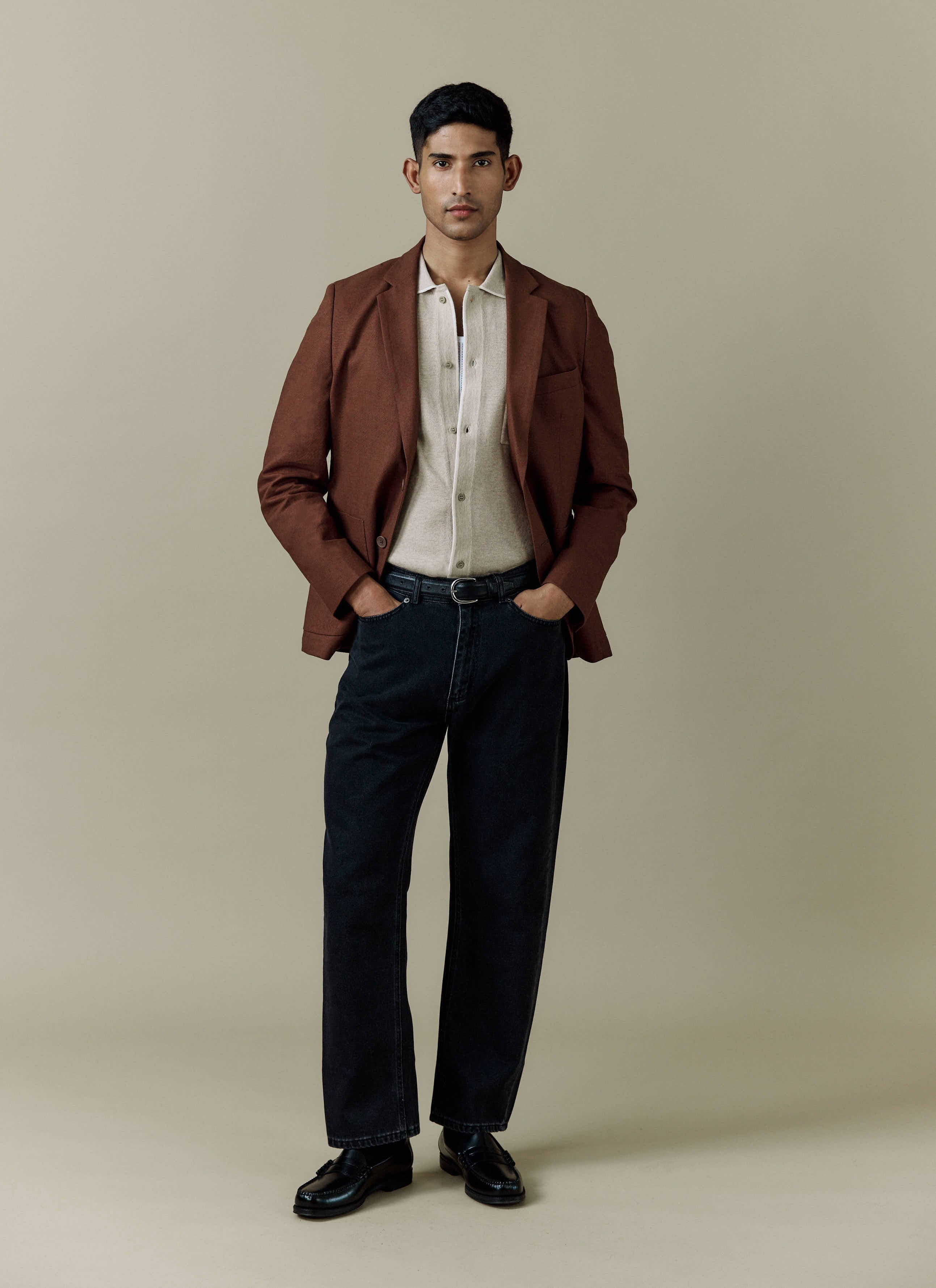 A model wears the Percival Tailored Linen Blazer in brown, styled with a beige knit shirt and dark jeans.