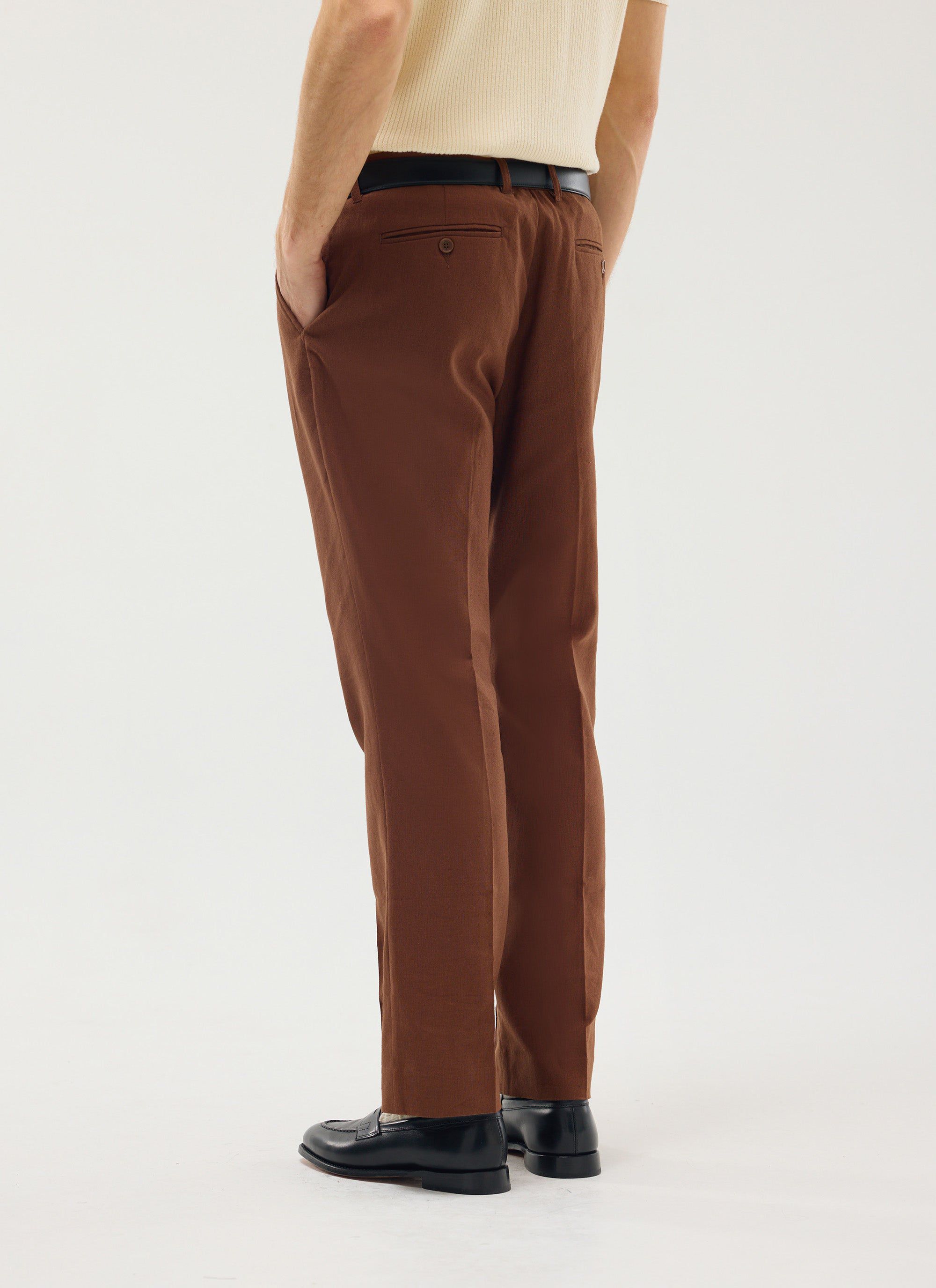A model wears the brown Tailored Linen Trousers by Percival, shown from behind with a cream knit top and black loafers.