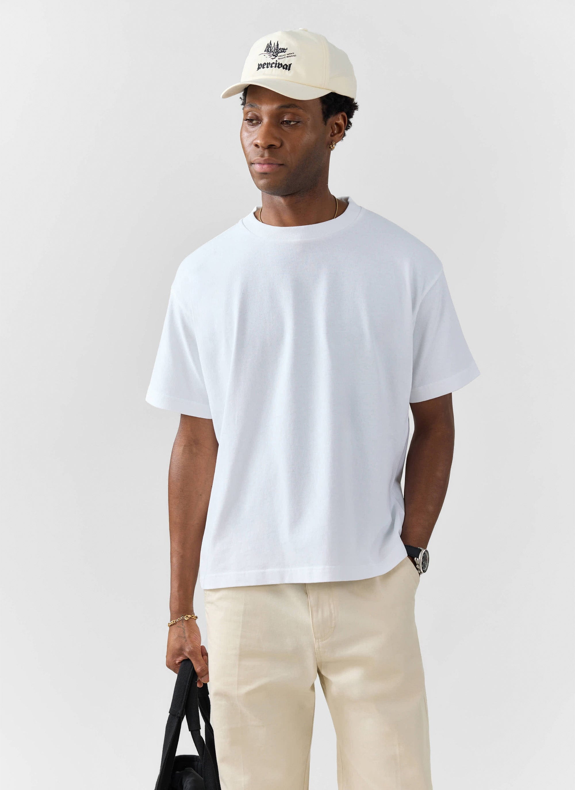 A model wears a Percival oversized white t-shirt with beige trousers and a cream Percival cap against a white background.