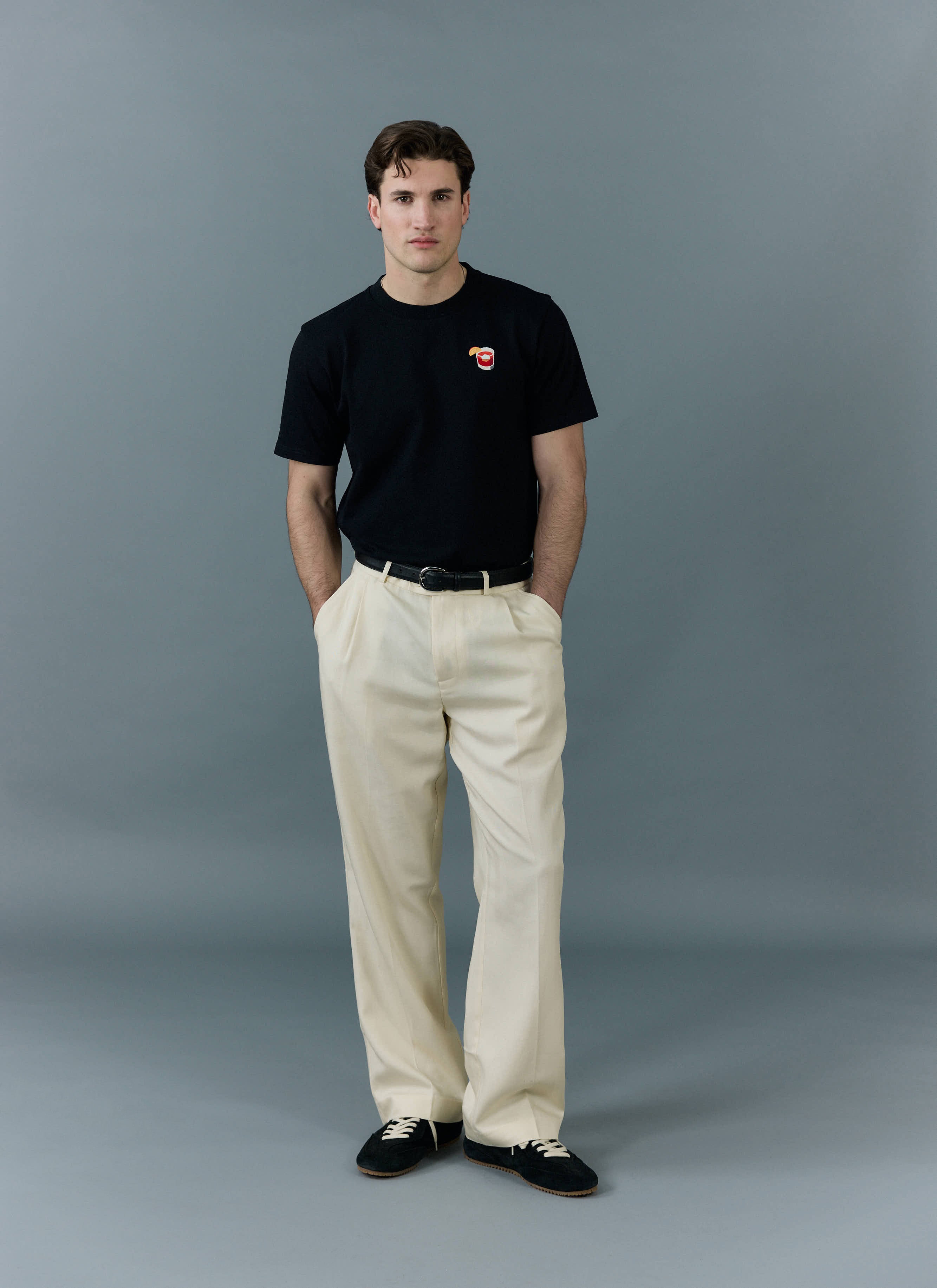 A model wears the black Negroni T-Shirt from Percival, tucked into smart cream trousers in a full-length studio shot.