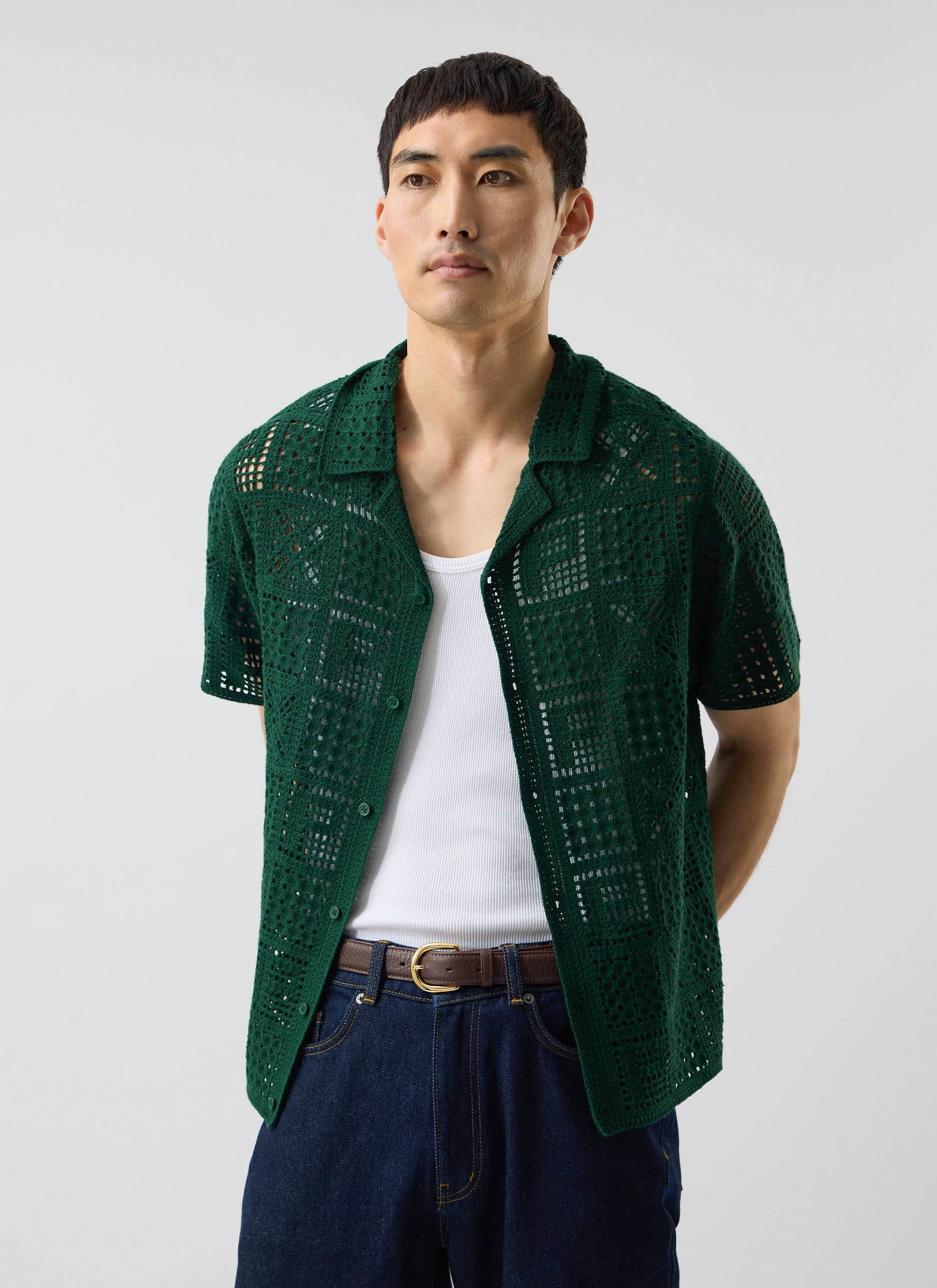 A model wears the Percival Crochet Groundsman Shirt in forest green, layered over a white vest and styled with dark jeans.