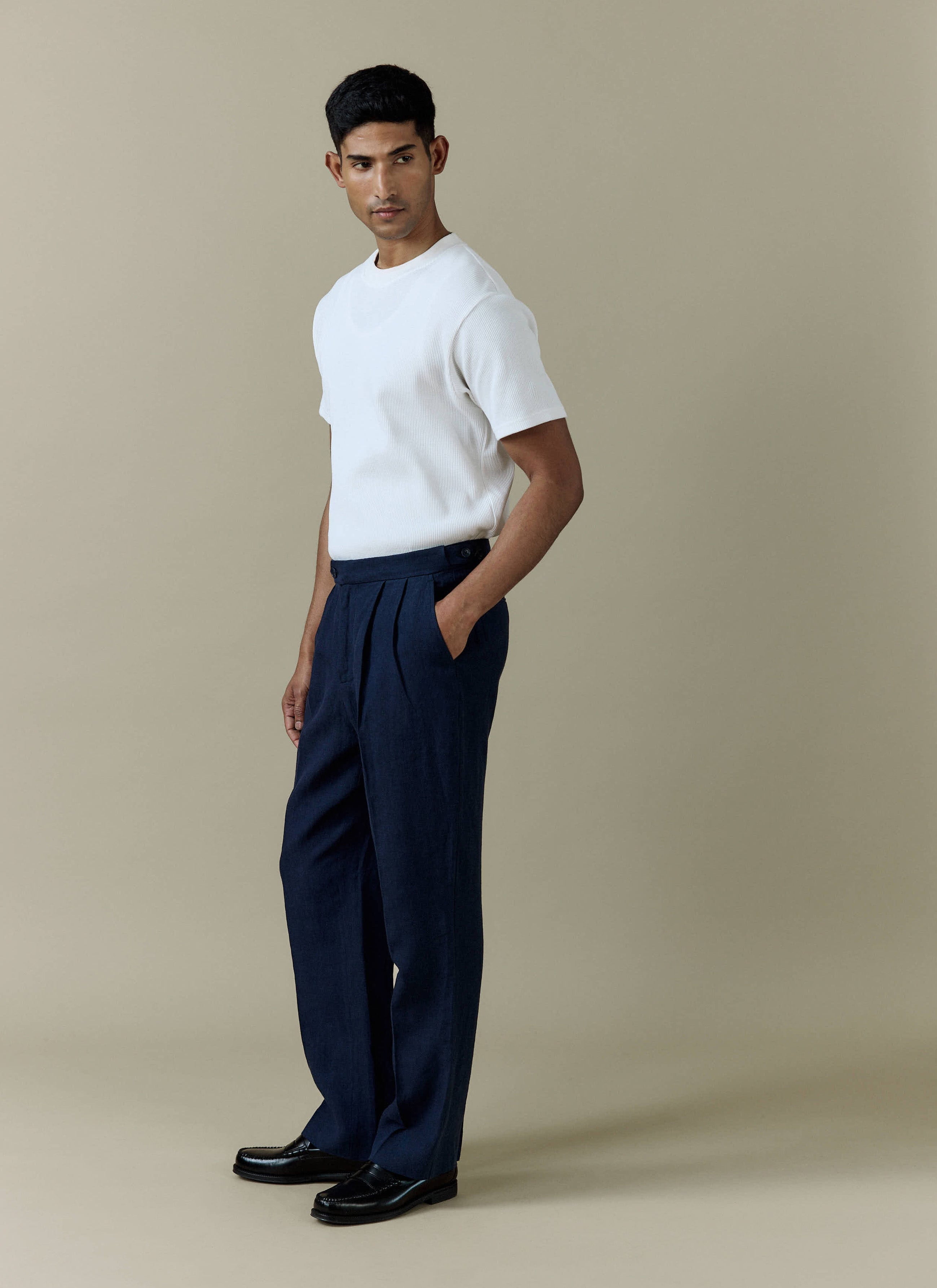 A model wears the Percival Pleated Tailored Trousers in navy linen, styled with a white t-shirt and black loafers.