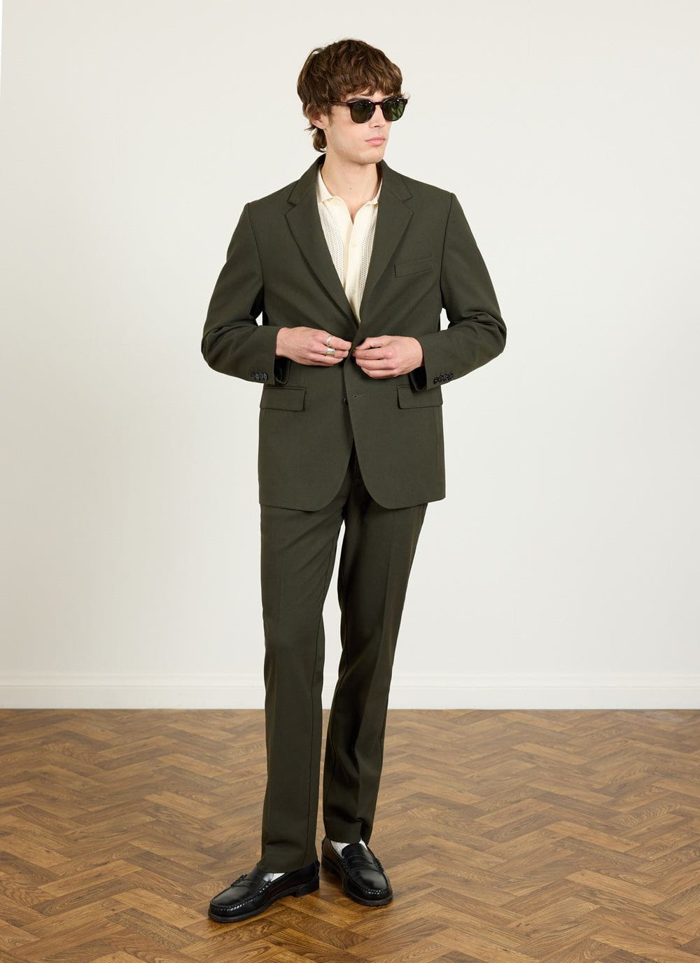 A model wears the Percival Tailored Blazer in forest green wool with matching trousers, a cream polo and sunglasses.