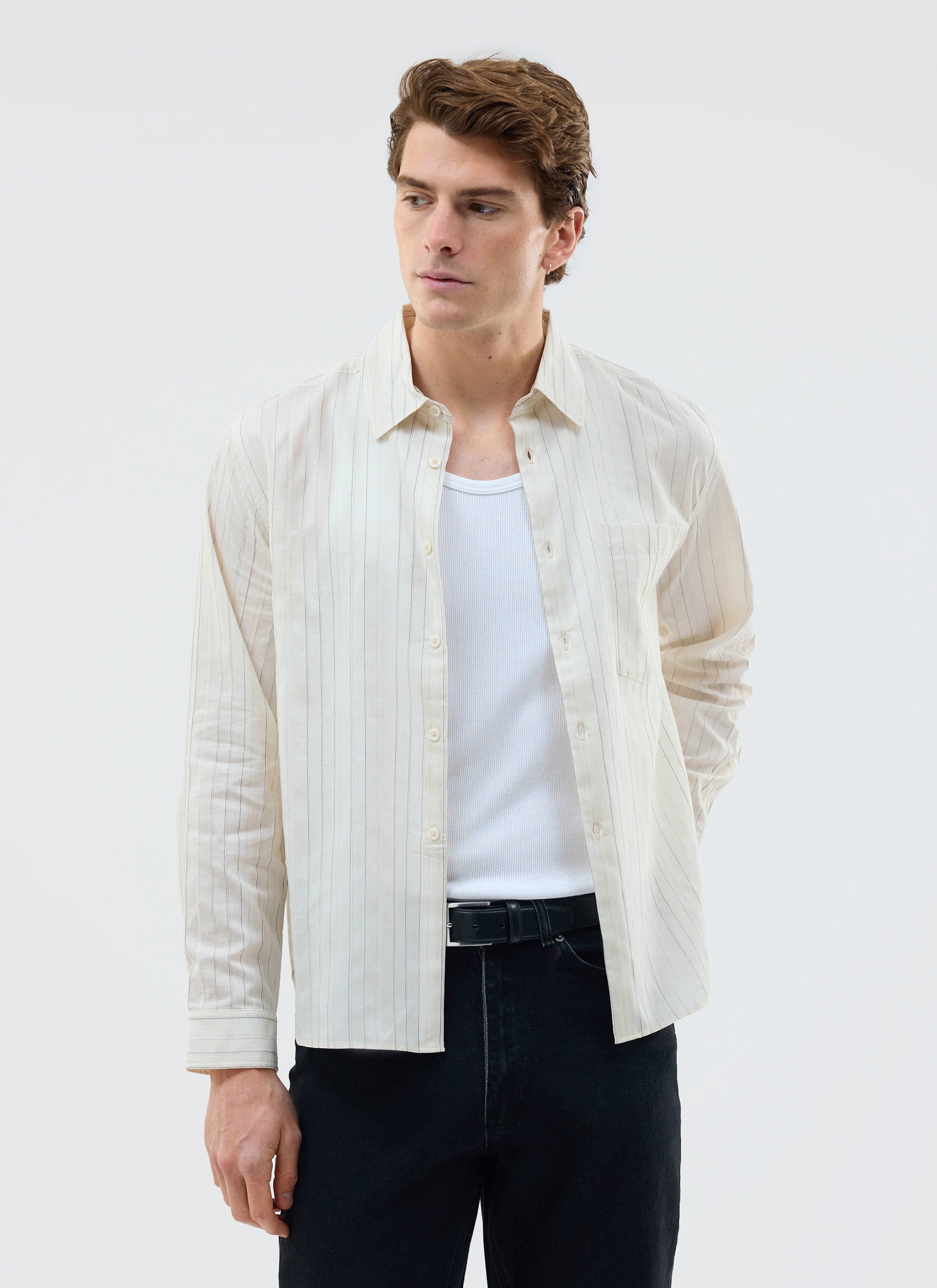 A man models the Percival Lorenzo Pearce Shirt in ecru, open over a white vest and black jeans against a plain background.