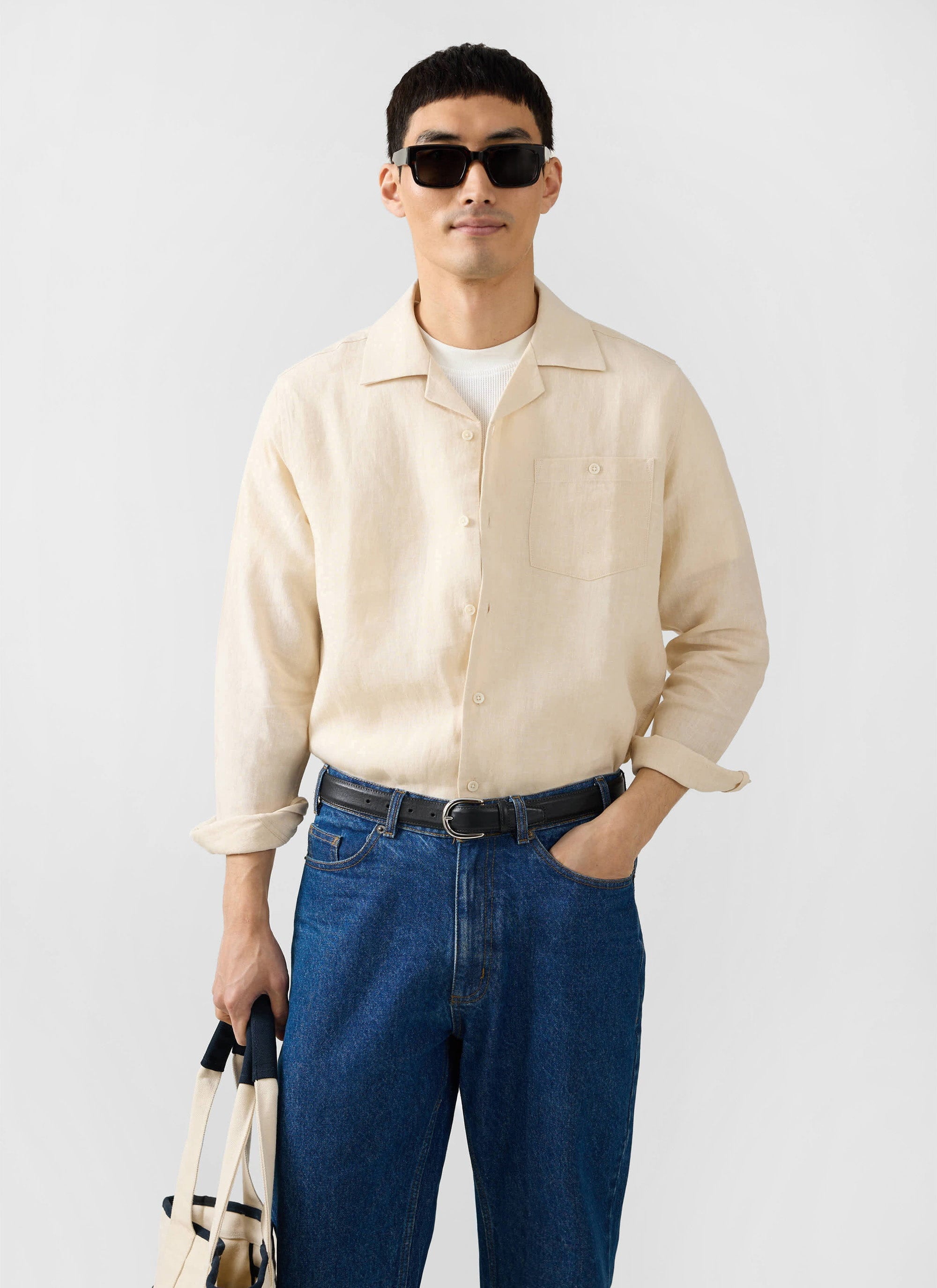 A model wears the Percival cream long sleeve Cuban linen shirt with blue jeans and sunglasses against a white background.