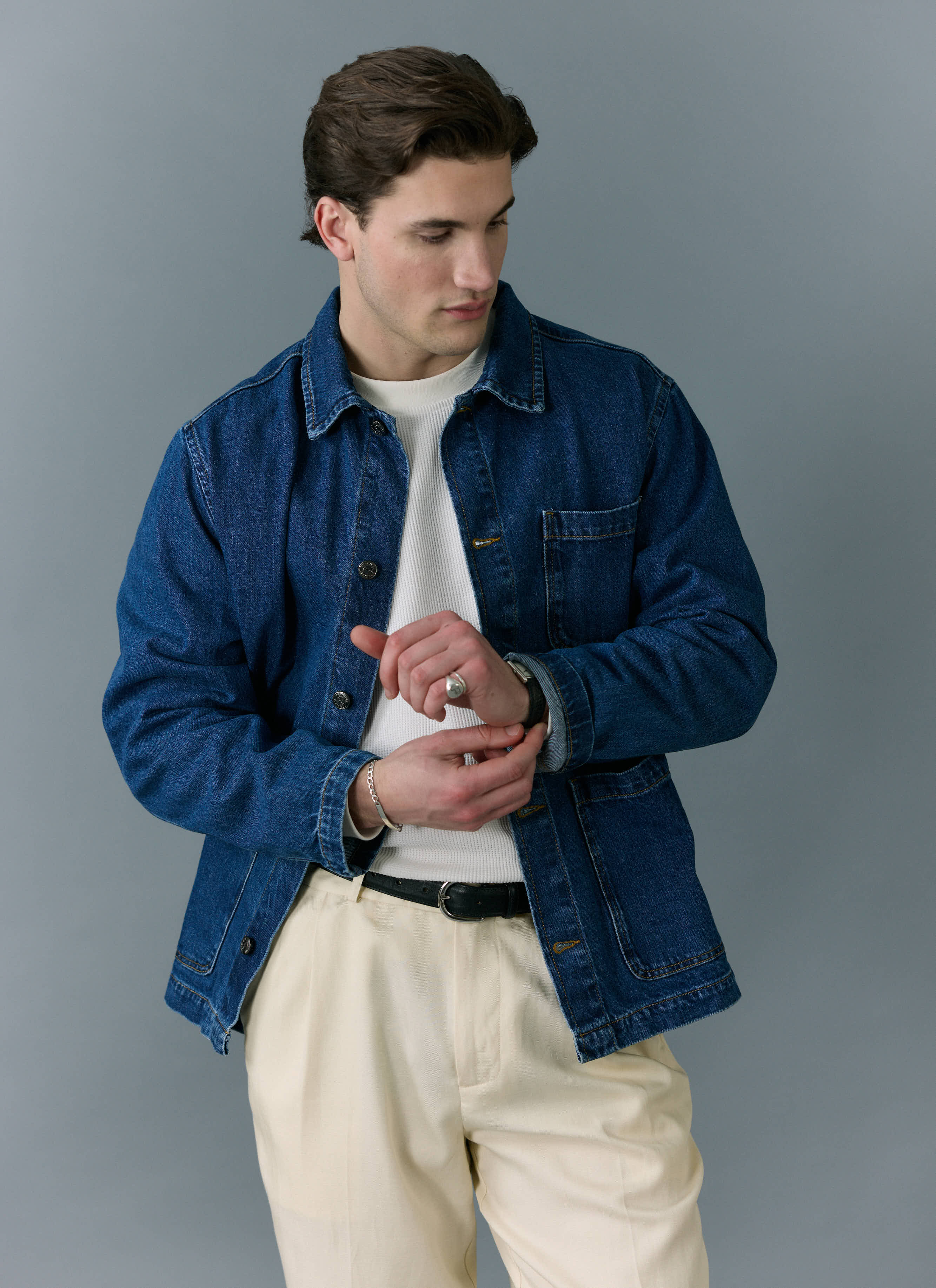 A model wears the Percival Gove Chore Jacket in mid-wash blue denim, adjusting his cuff against a grey background.