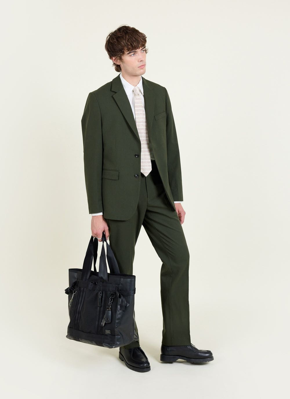 A model wears the Percival forest green wool tailored blazer with matching trousers, a white shirt, tie, and a black bag.