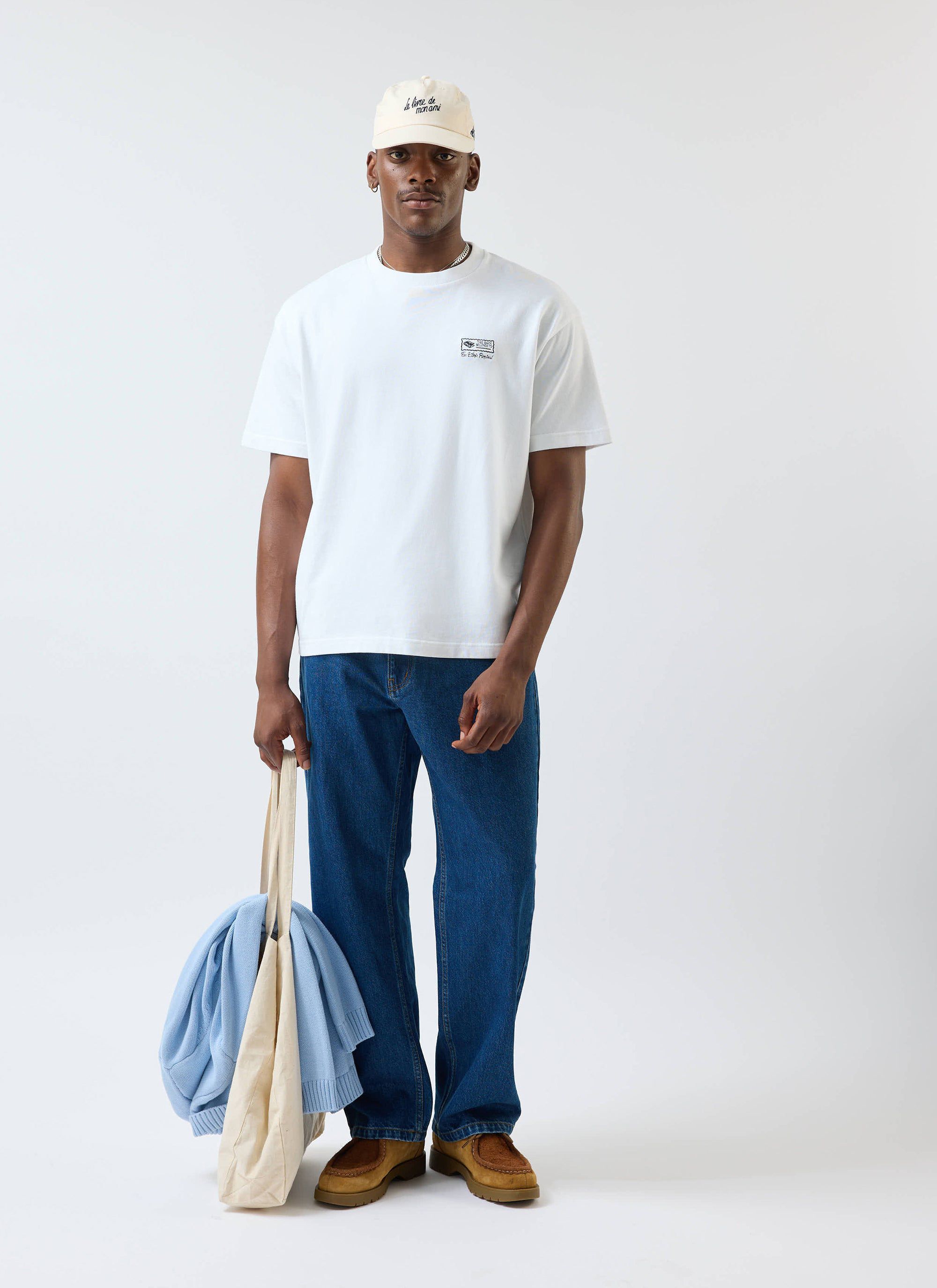 A model wears the white Percival Ex Libris t-shirt, paired with blue jeans, a cream cap and holding a tote bag.