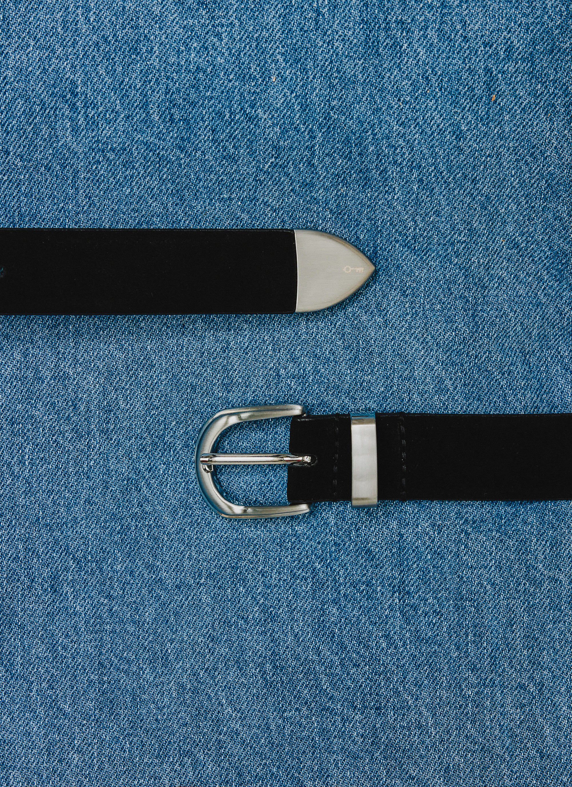 The Percival Suede Belt in black leather, with silver buckle and western tip, laid out across a blue denim background.