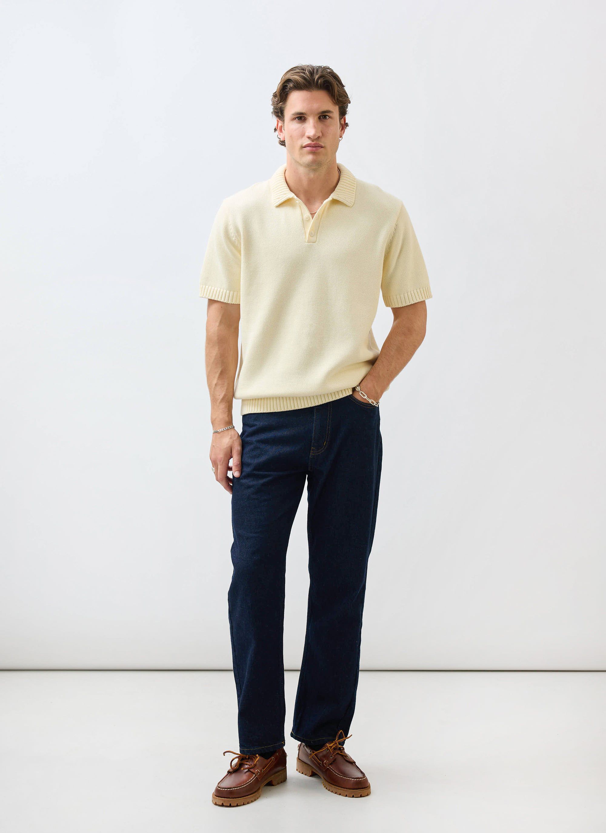 A model wears the Percival Verso Short Sleeve Knitted Shirt in ecru, styled with dark denim jeans and brown leather shoes.