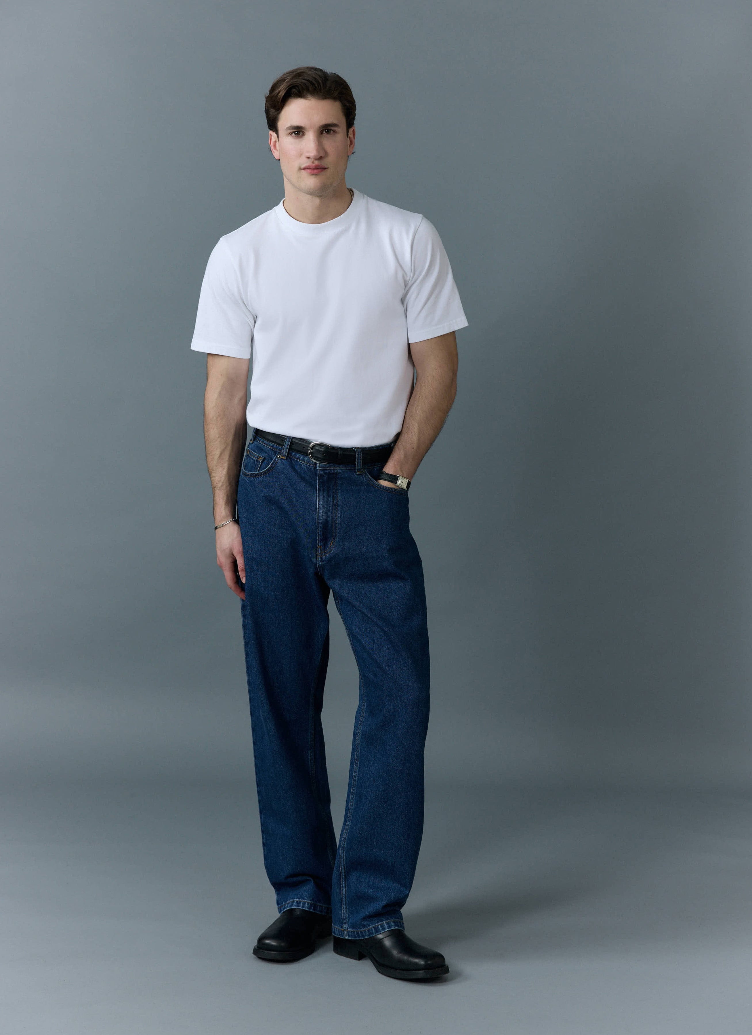 A man models the Percival Wide Leg Denim Jeans in mid wash blue with a white t-shirt against a grey background.