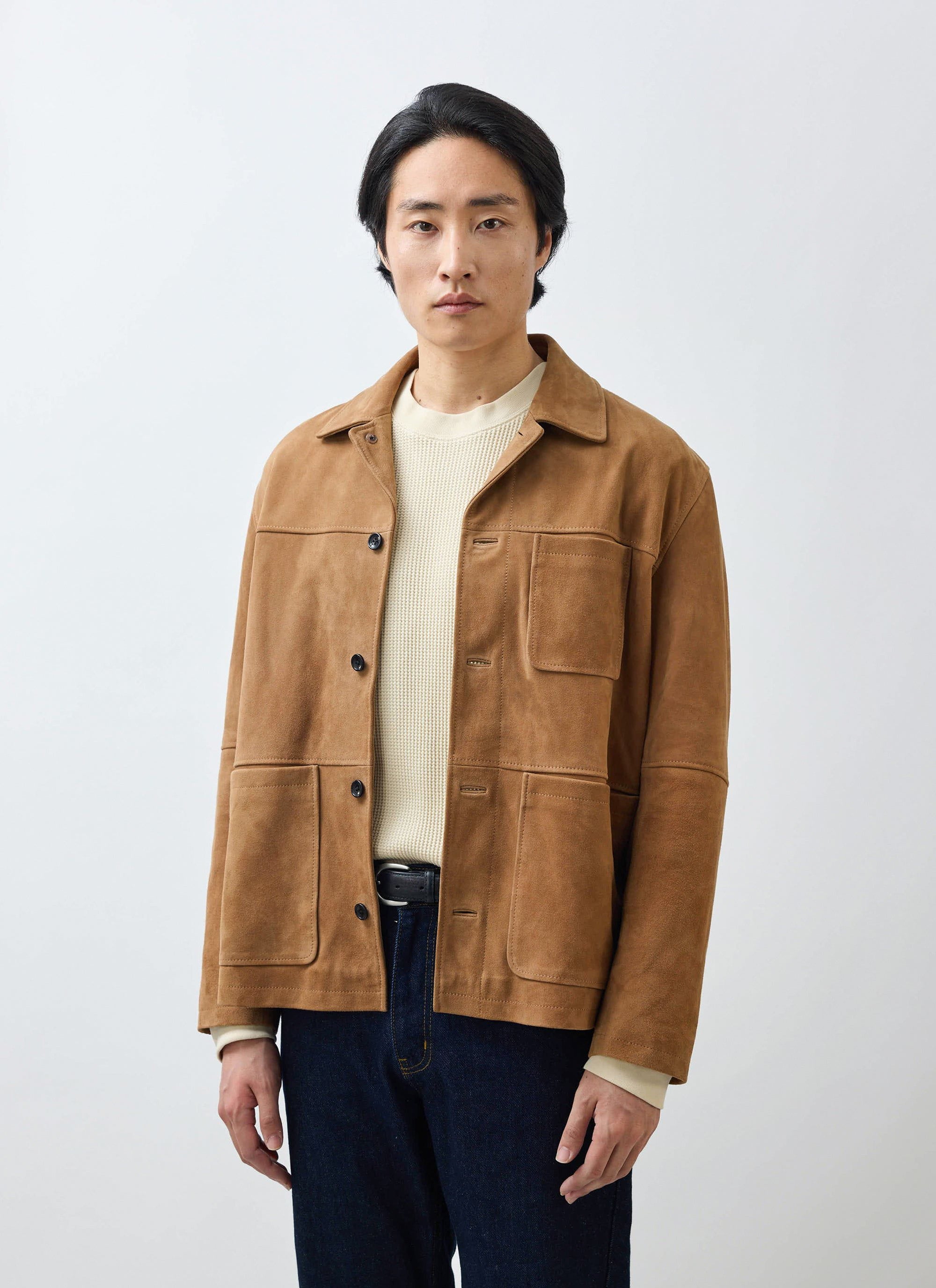 A model wears the tan Afterward Suede Chore Jacket from Percival, styled with a cream knit jumper and dark denim.