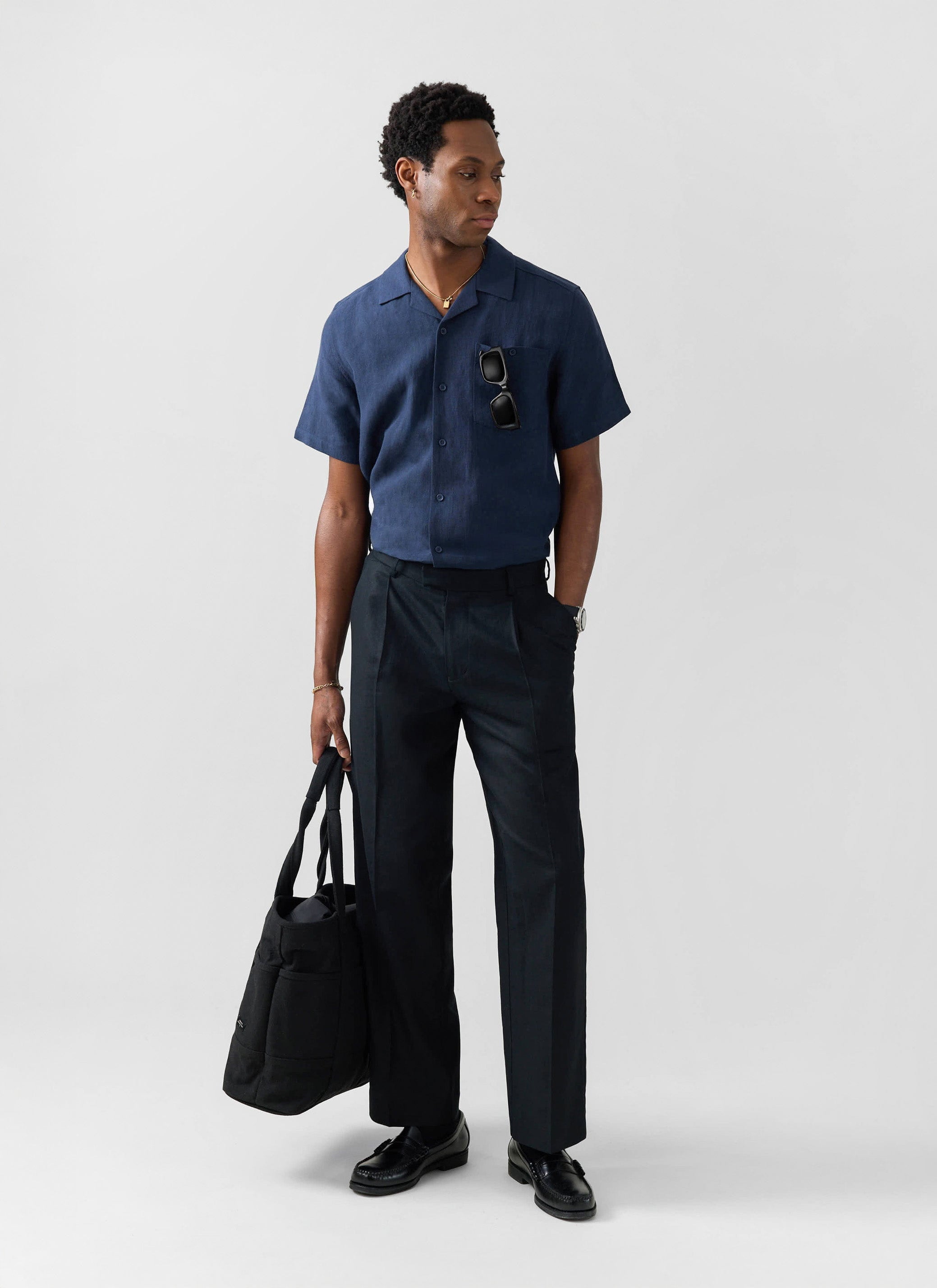 A model wears the Percival navy blue linen short sleeve Cuban shirt with black trousers, holding a black tote bag.