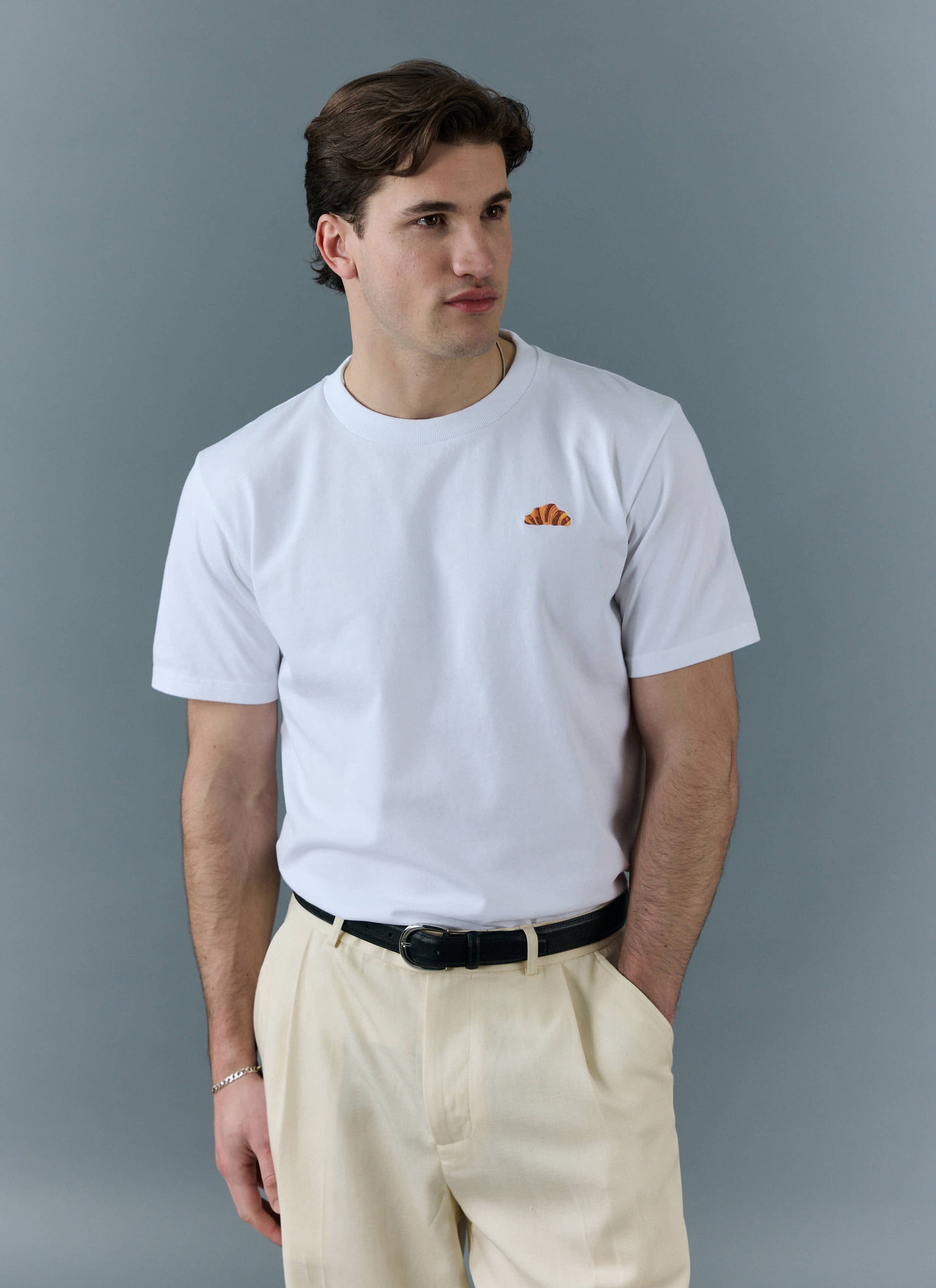 A model wears the Percival white t-shirt with an embroidered croissant on the chest, styled with cream trousers.