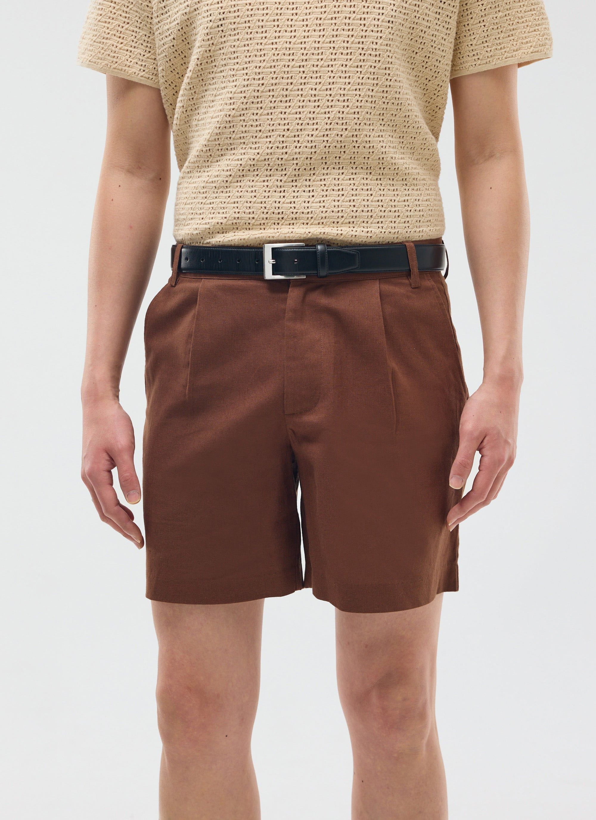 A model wears the Percival Pleated Linen Shorts in brown, styled with a beige knitted t-shirt and a black leather belt.