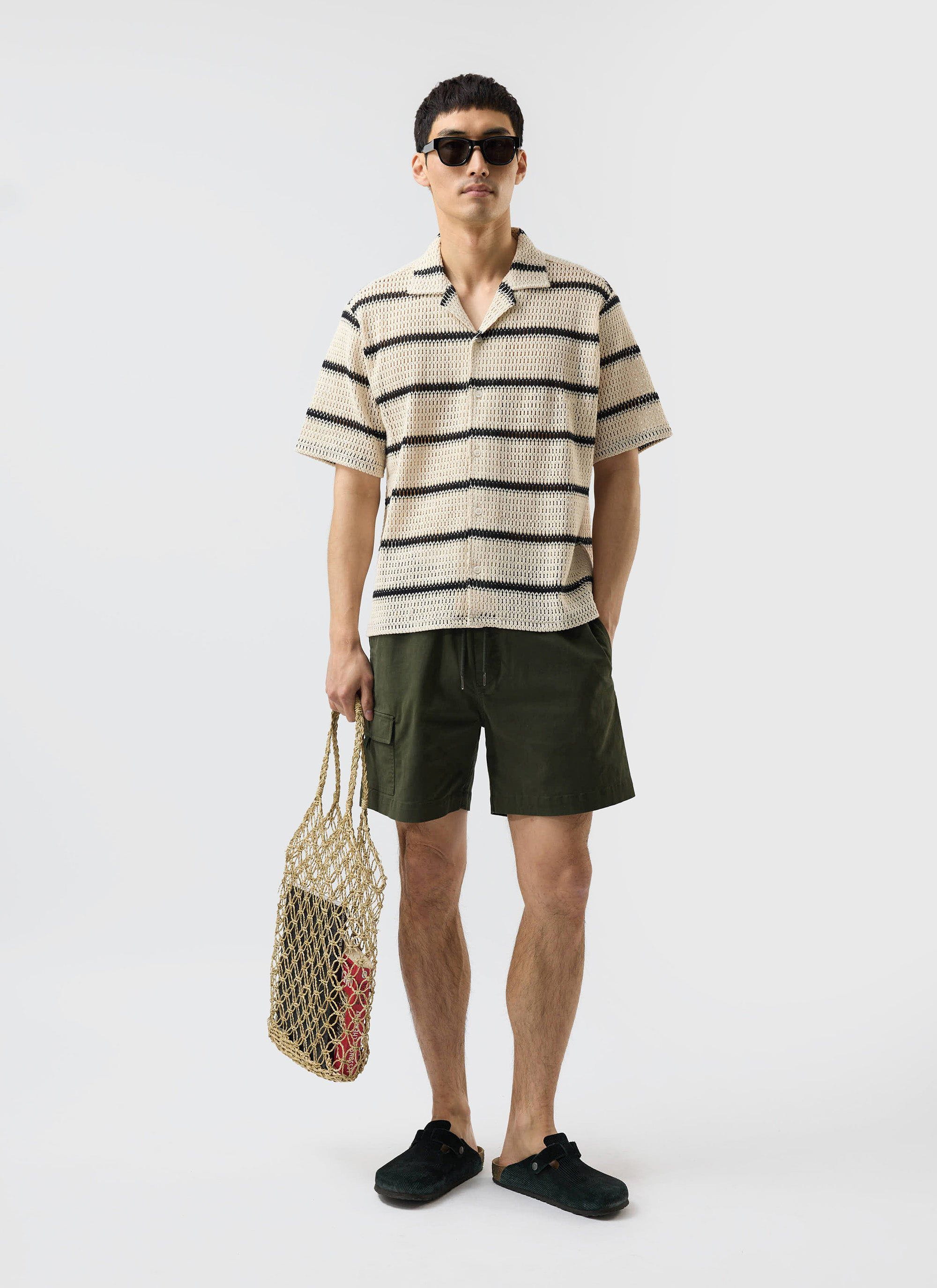 A man models the ecru and black striped Crochet Groundsman Cuban Shirt from Percival with green shorts and sunglasses.