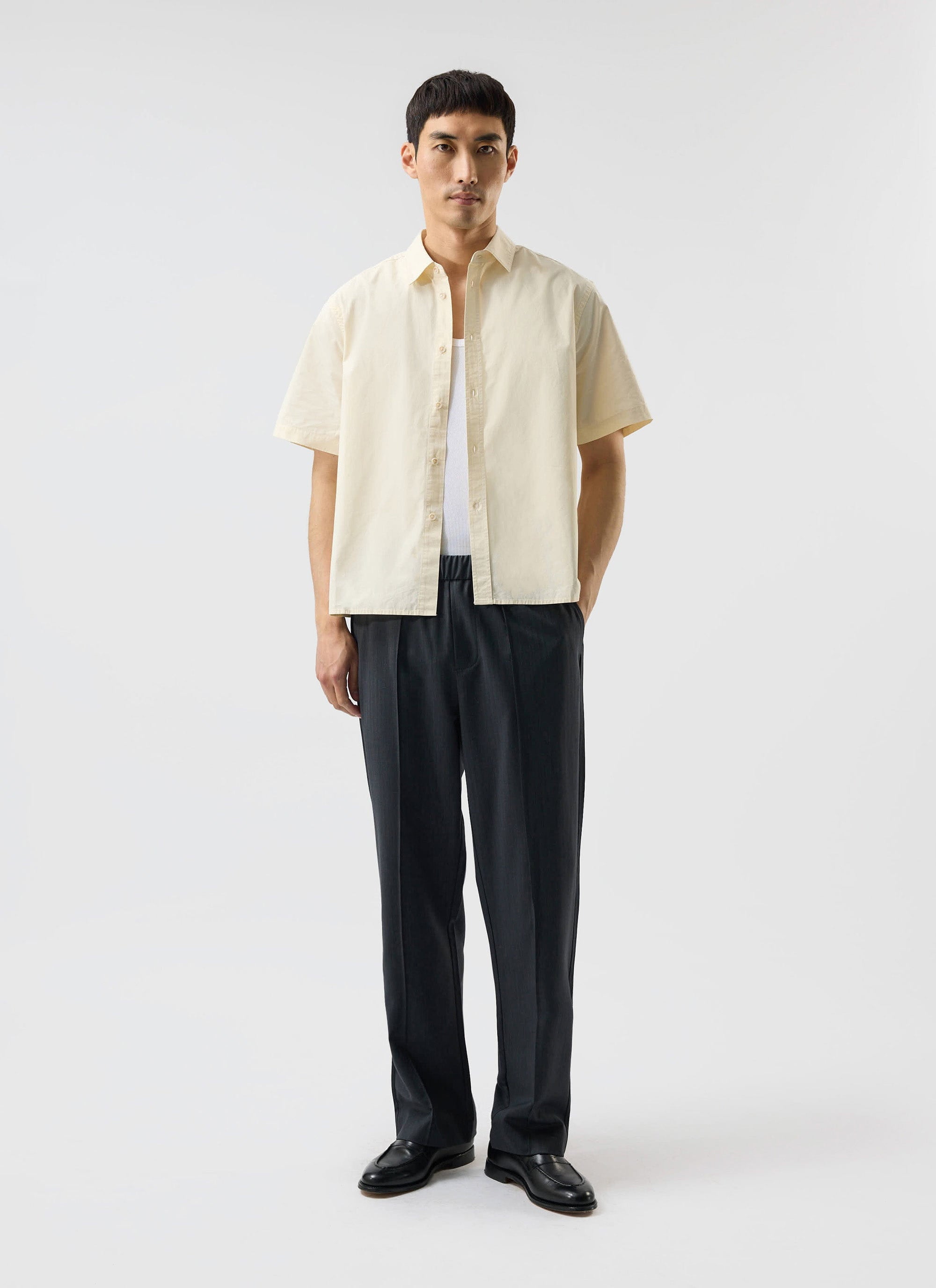 A model wears the Percival Boxy Cropped Clerk Shirt in ecru, styled open over a white vest and dark trousers.