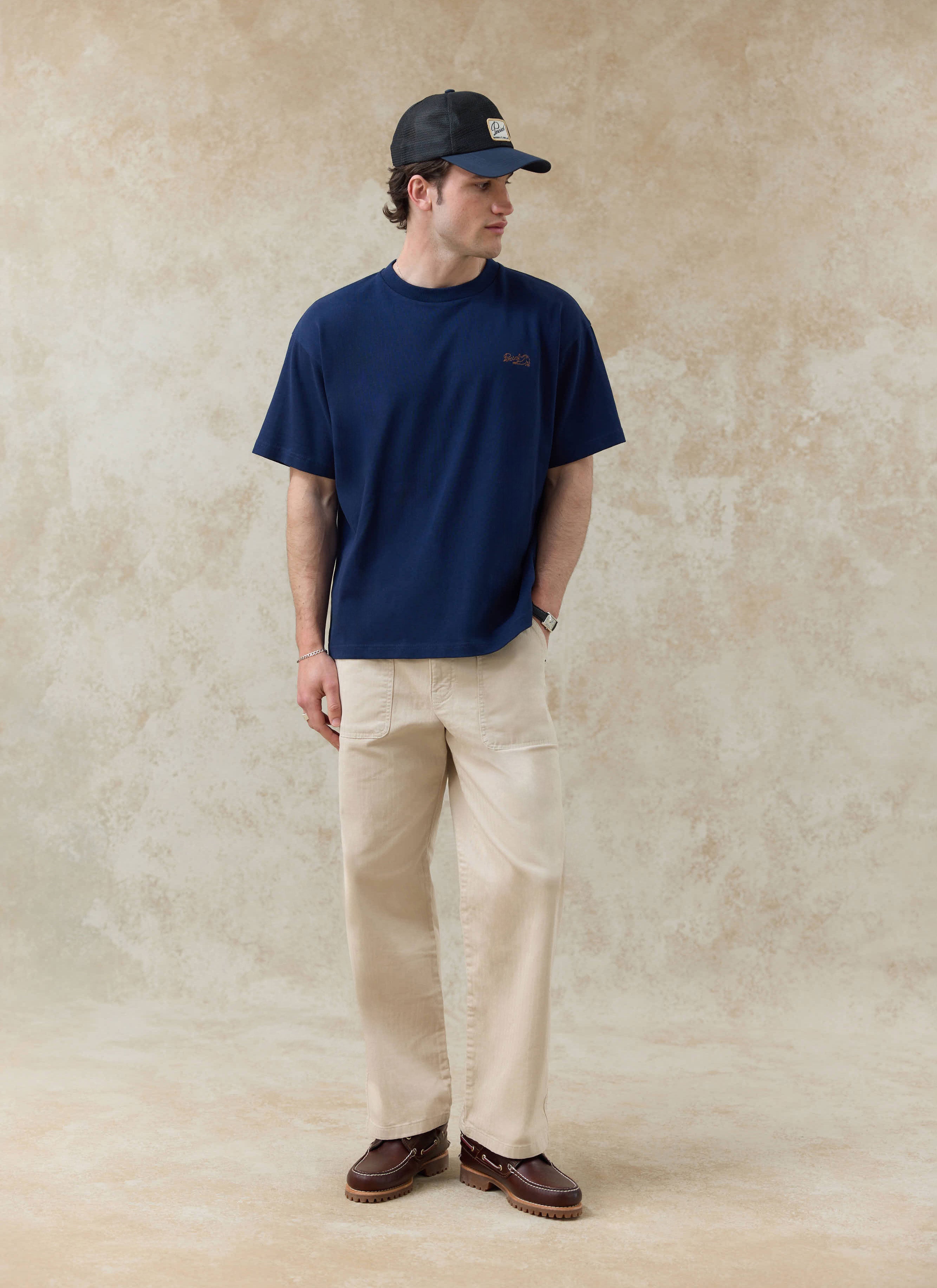 The navy Percival Clarke Embroidered T-shirt in organic cotton, styled with beige trousers and a mesh cap for SS26.