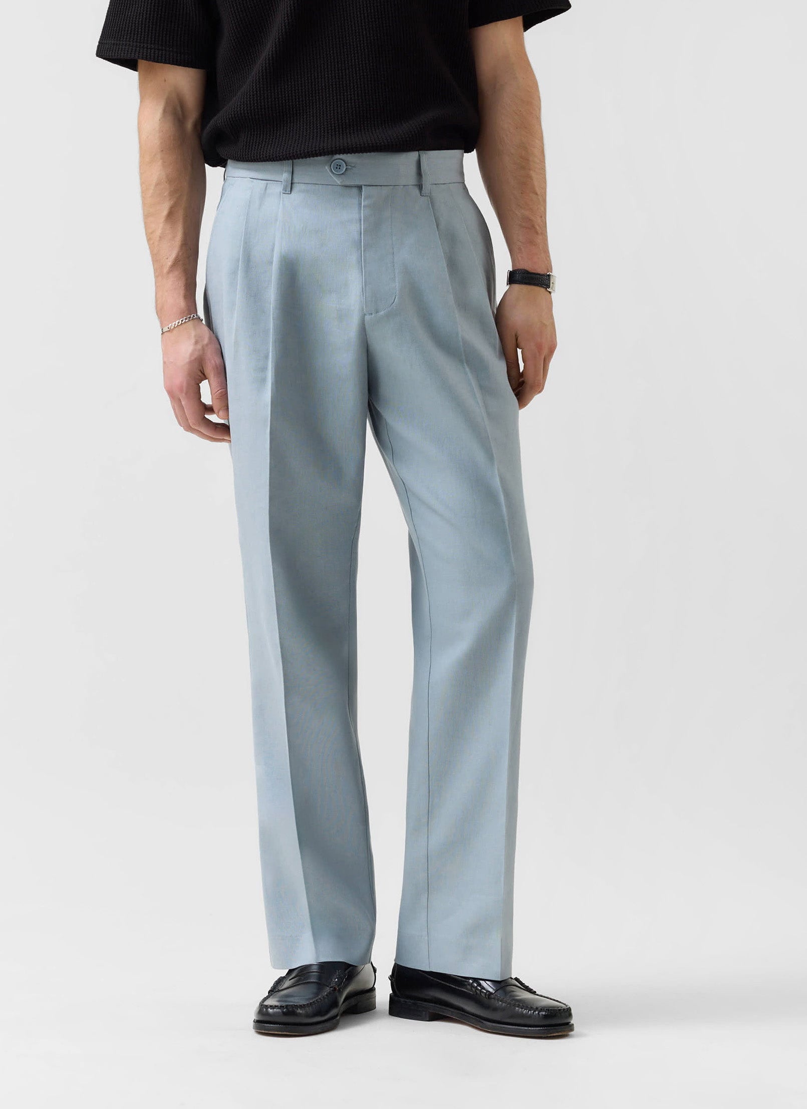 A model wears the Percival Pleated Tailored Trousers in light blue linen with a black top and black loafers.