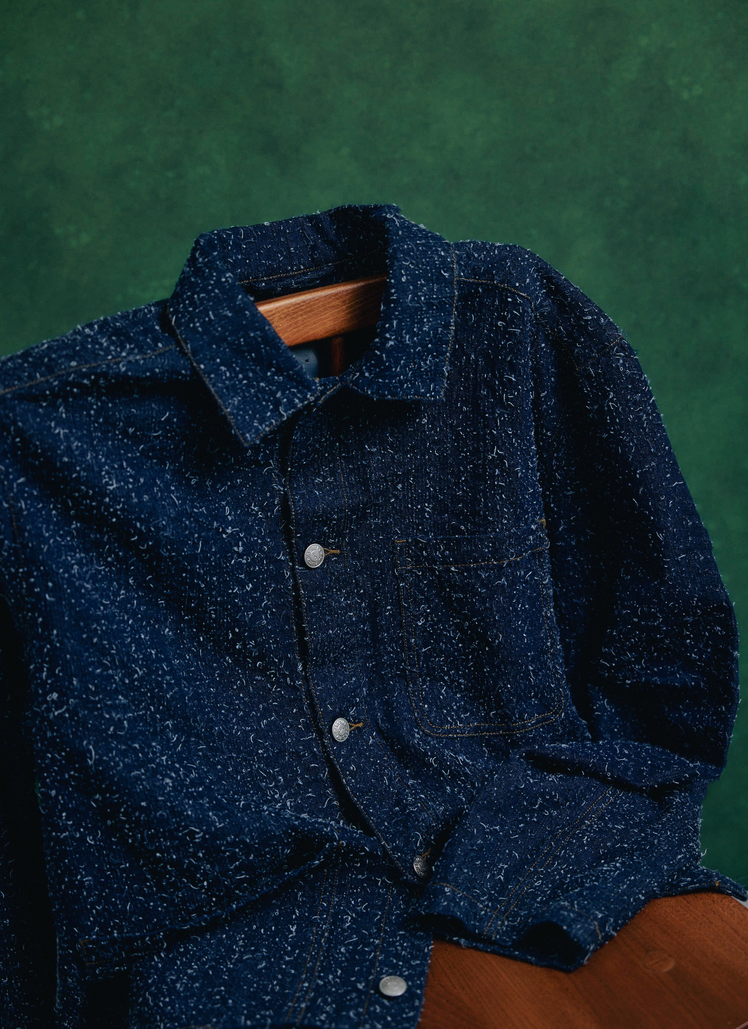 Navy blue textured nep cotton Percival Parsons Chore Jacket, draped over a wooden chair against a green background.