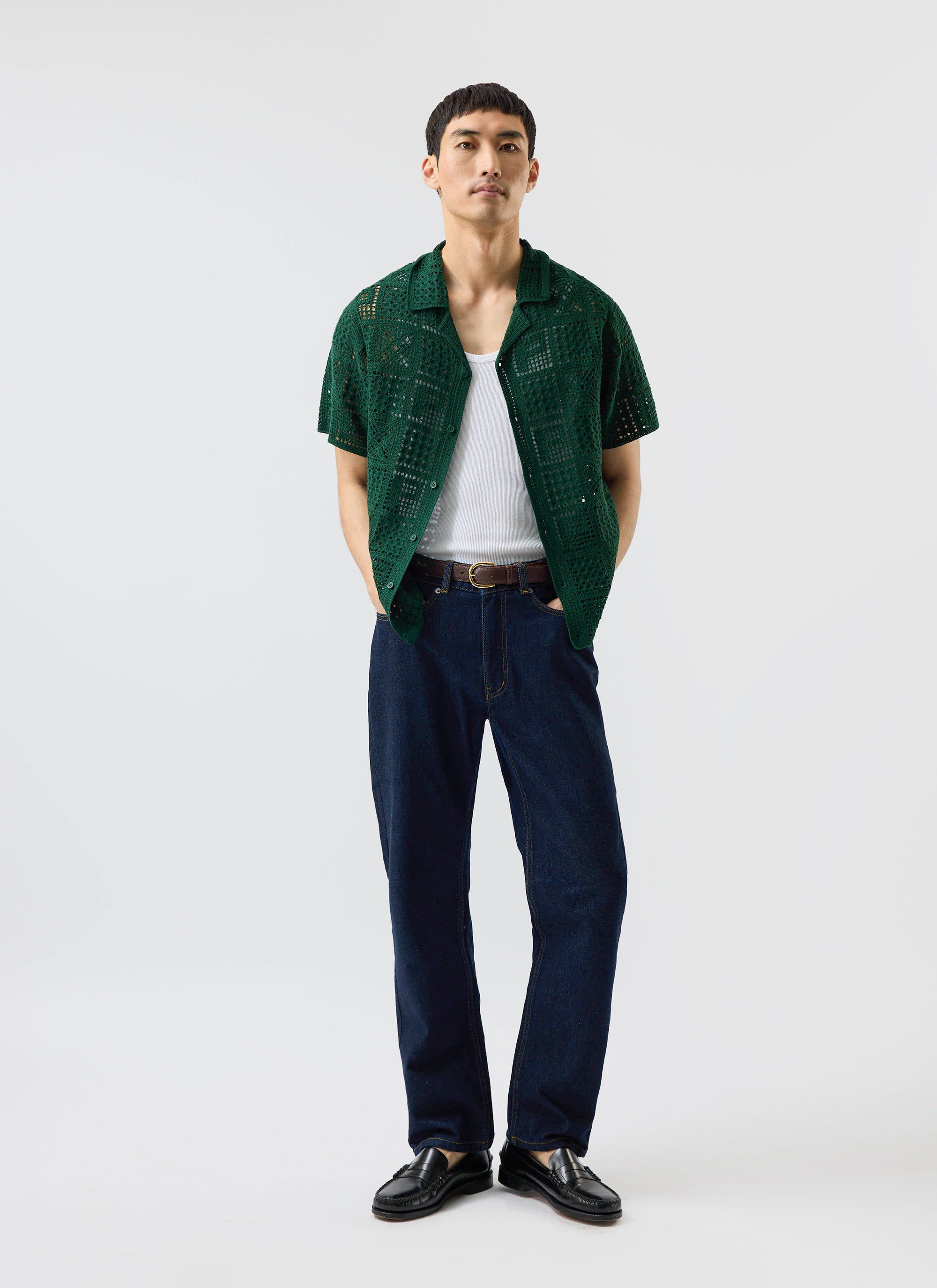 A man models the forest green Percival Crochet Groundsman Shirt, worn open over a white vest and styled with dark jeans.