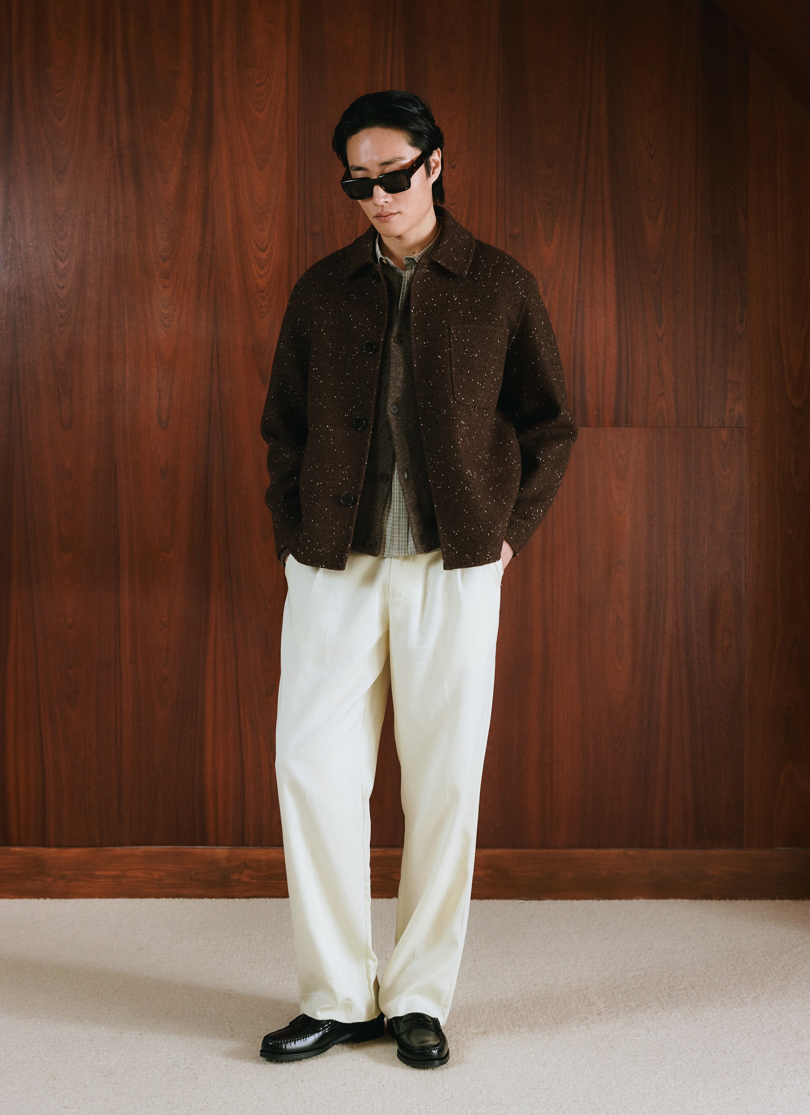 A model wears the Percival wool overshirt in brown fleck with cream trousers against a wood panel wall.