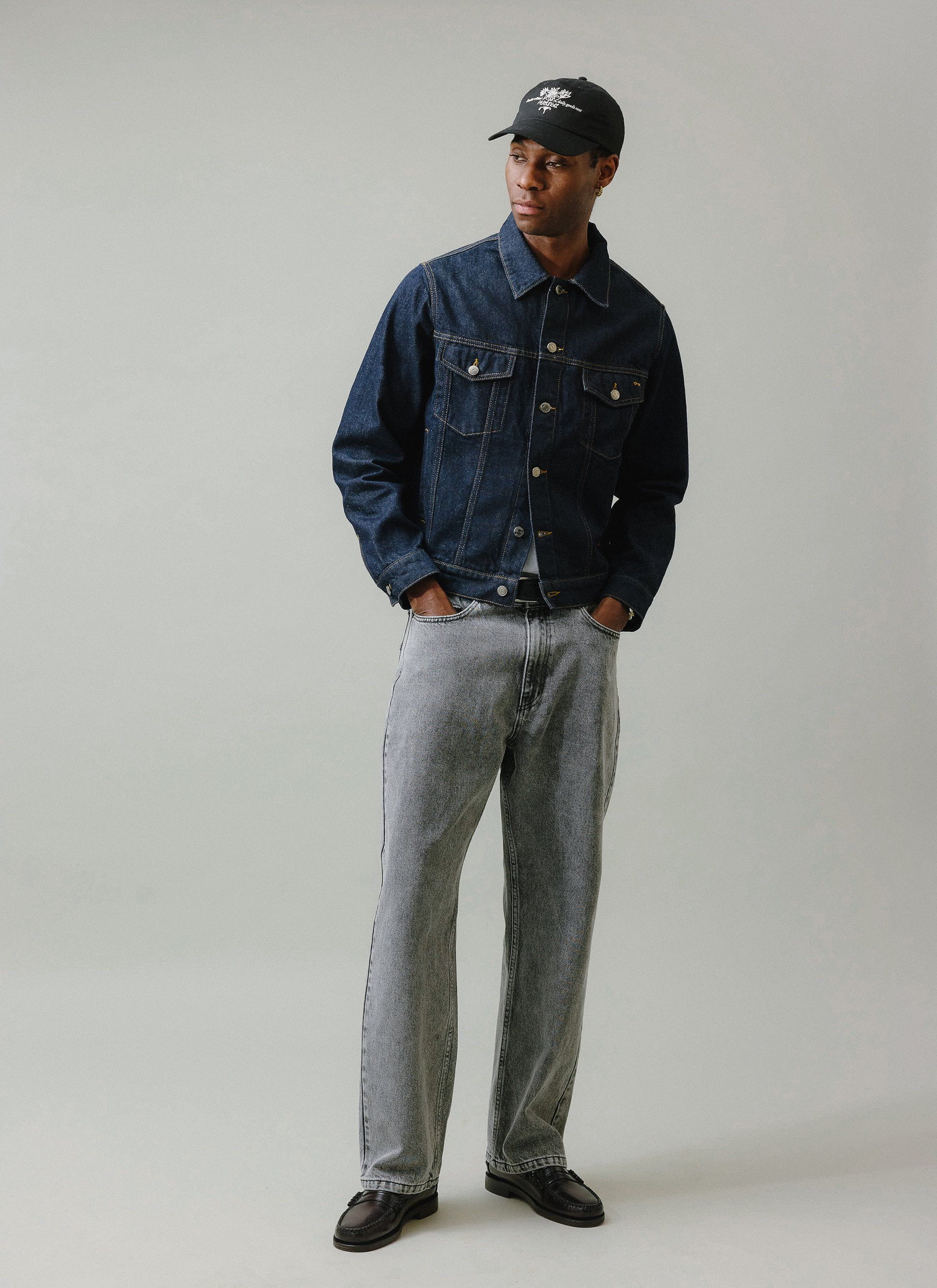 A model wears the Percival Wide Leg Denim Jeans in warm grey wash, styled with a blue denim jacket and a black cap.