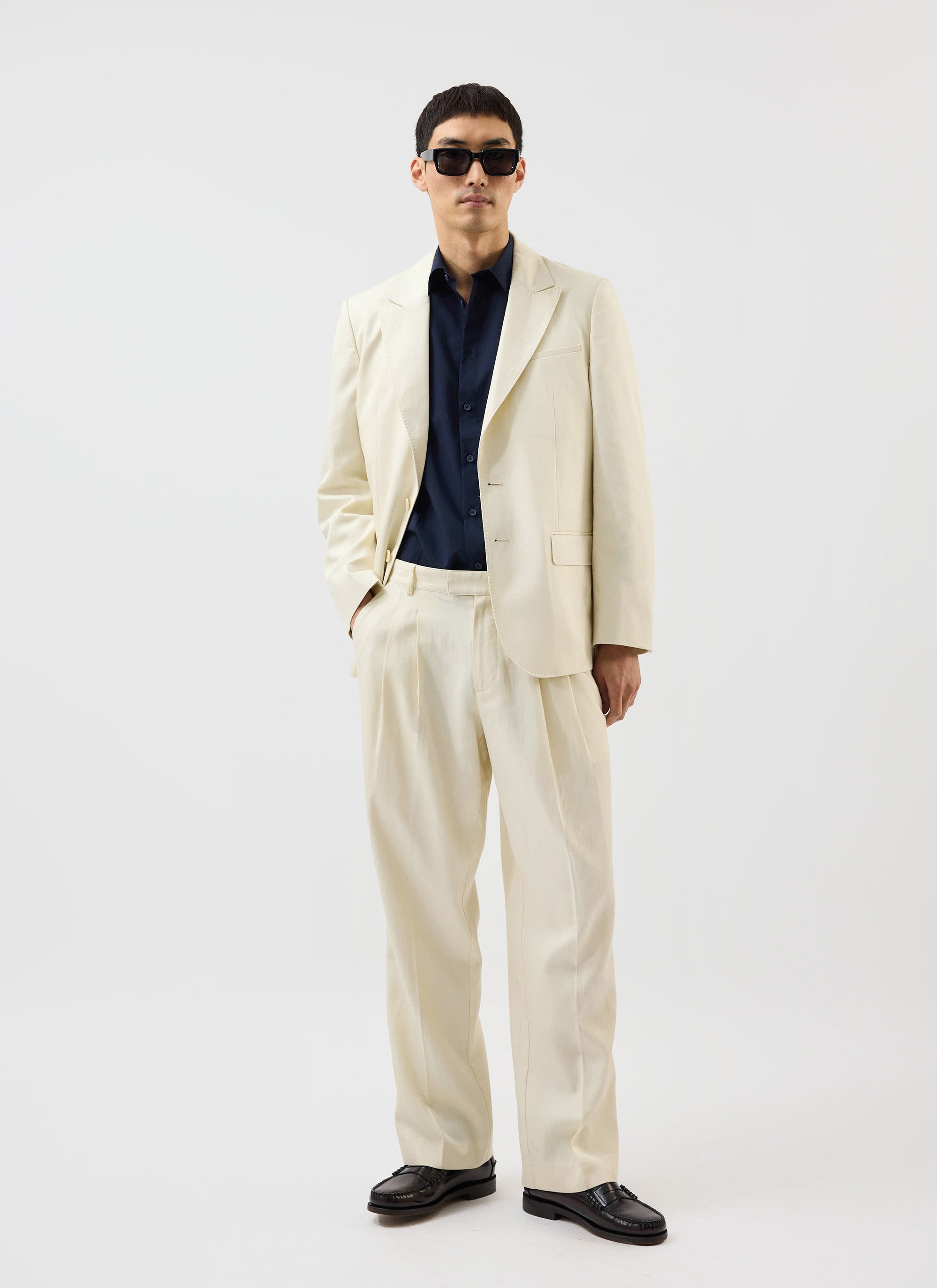 A model wears the Percival x Warren ecru tailored blazer with matching trousers, a navy shirt, and sunglasses.