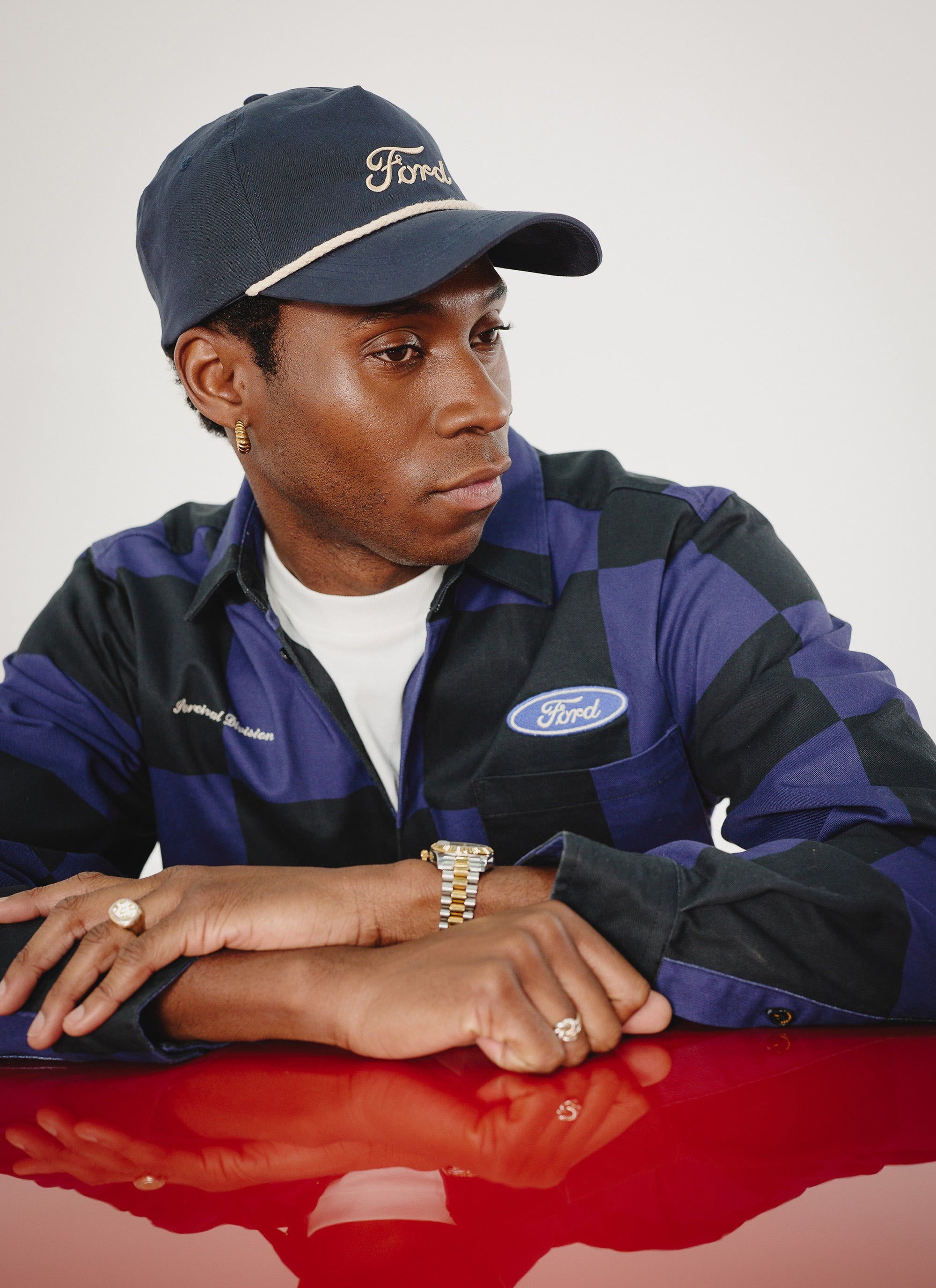 A model wears the Percival x Ford navy and black checkerboard overshirt and navy Ford cap while leaning on a red car bonnet.