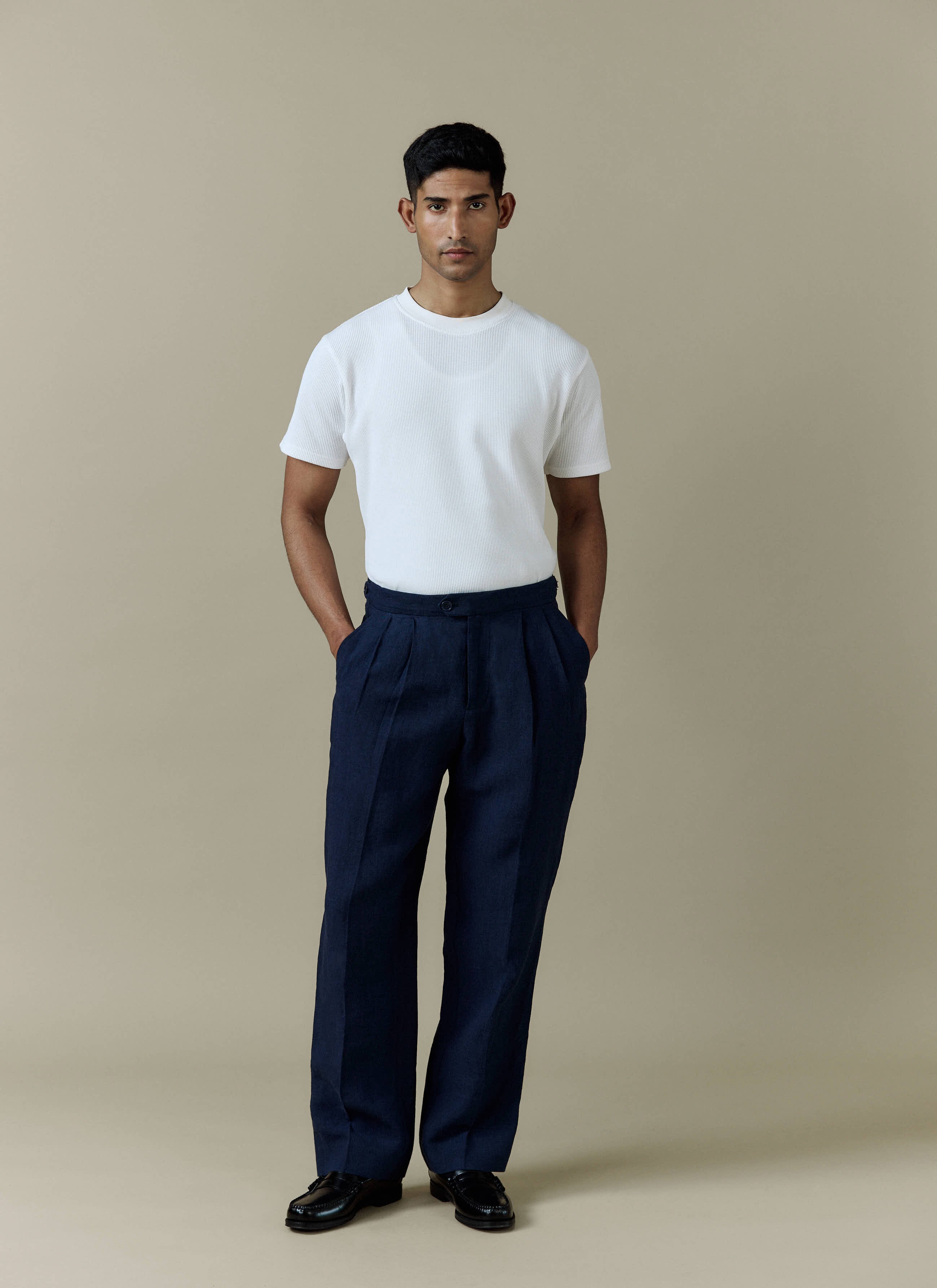 A man models the Percival Pleated Tailored Trousers in navy linen, styled with a white t-shirt and black loafers.