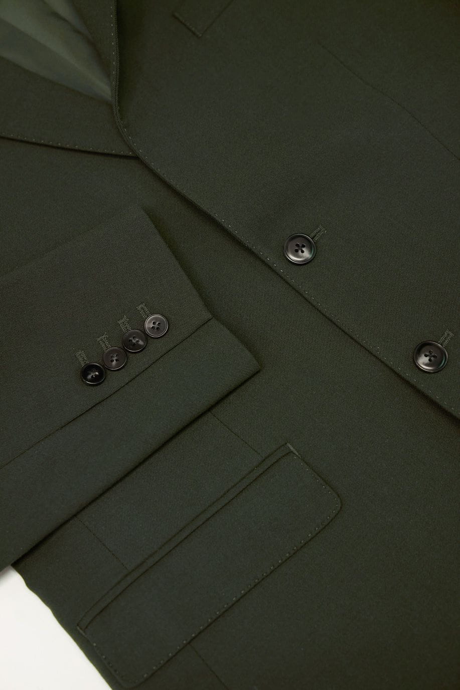 Close up of the Percival Tailored Blazer in forest green wool, showing the detail of the lapel, pocket, and buttoned cuff.