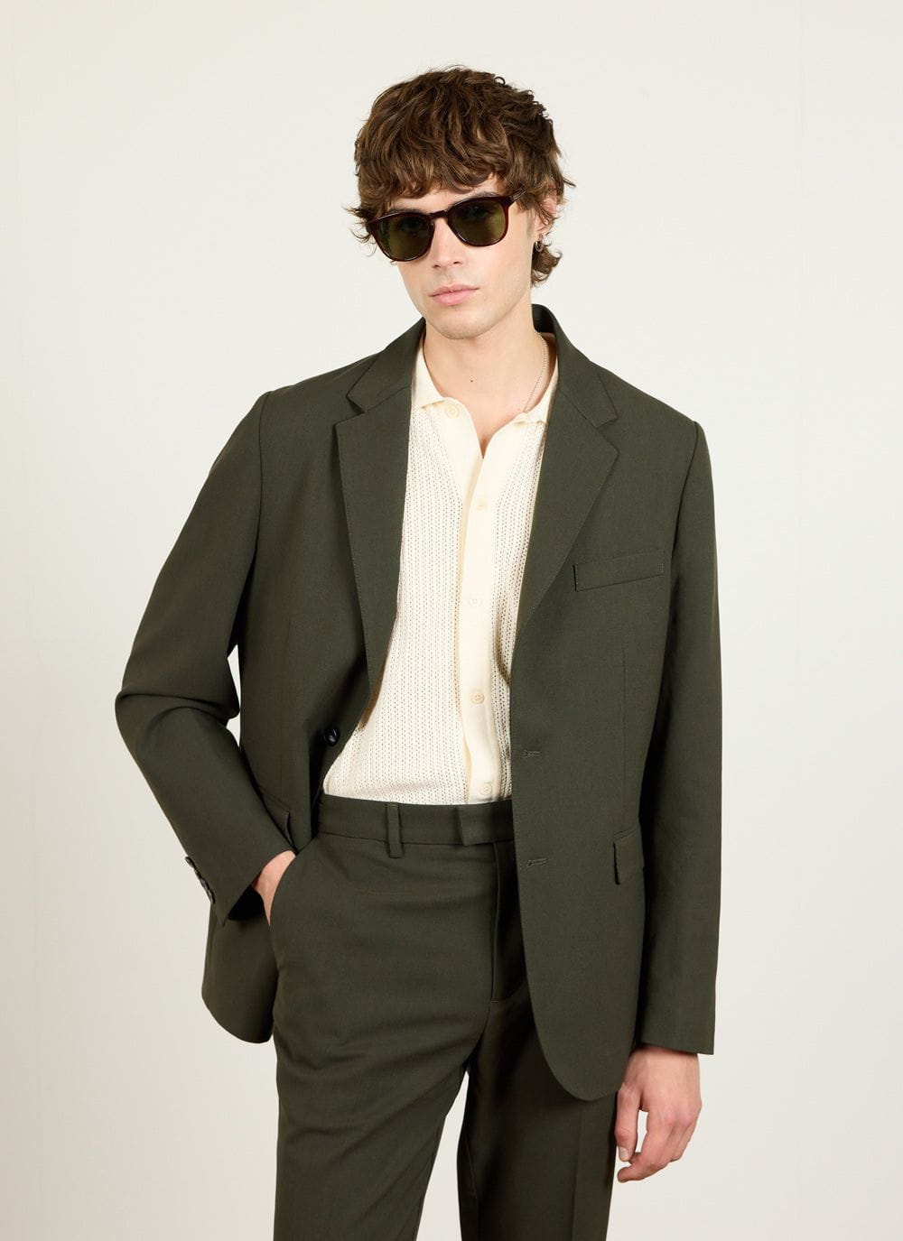 A model wears Percival's forest green wool tailored blazer and matching trousers with a cream knitted polo and sunglasses.