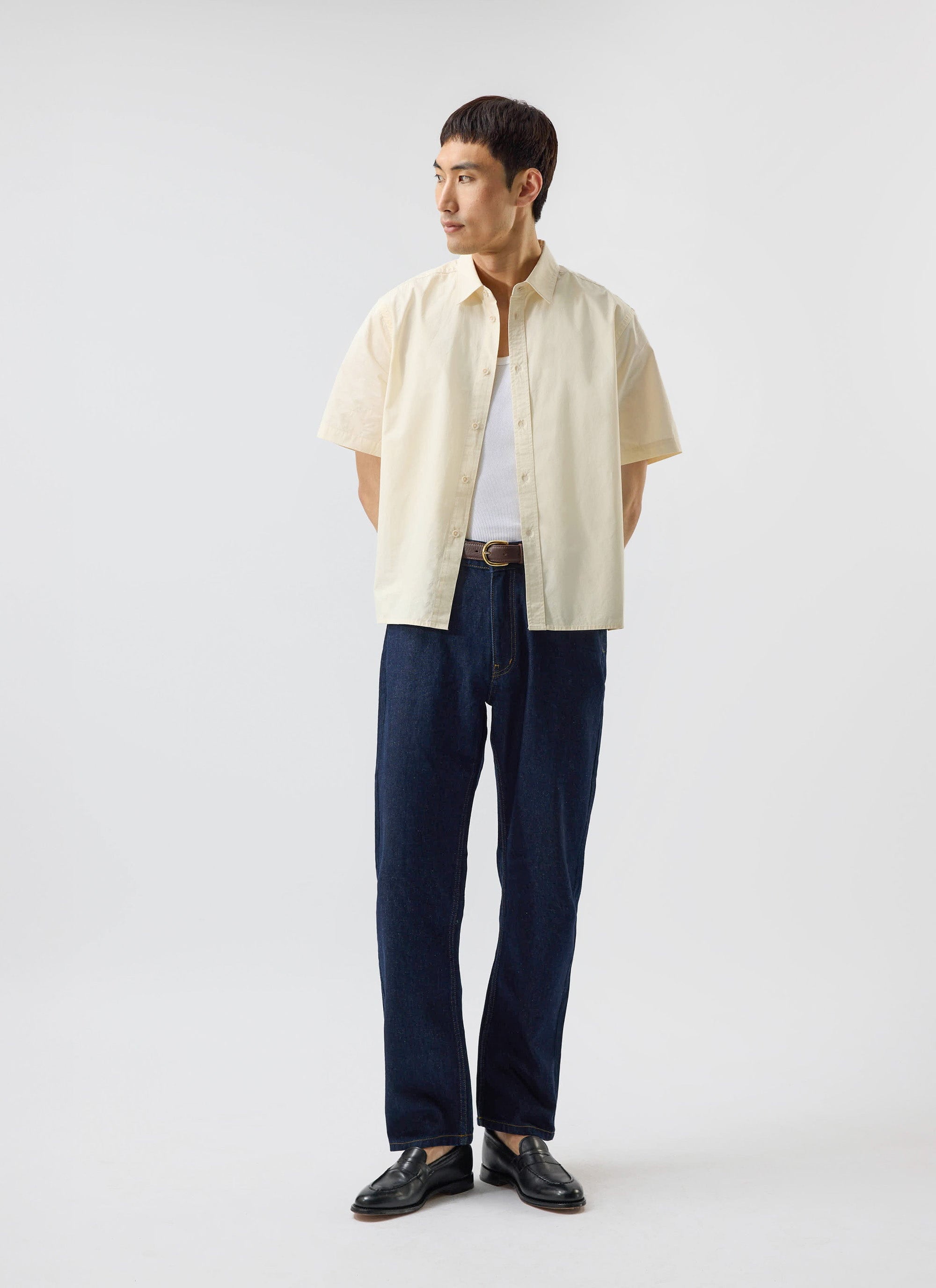 A man models the Percival Boxy Cropped Clerk Shirt in ecru, layered over a white vest and styled with dark denim jeans.