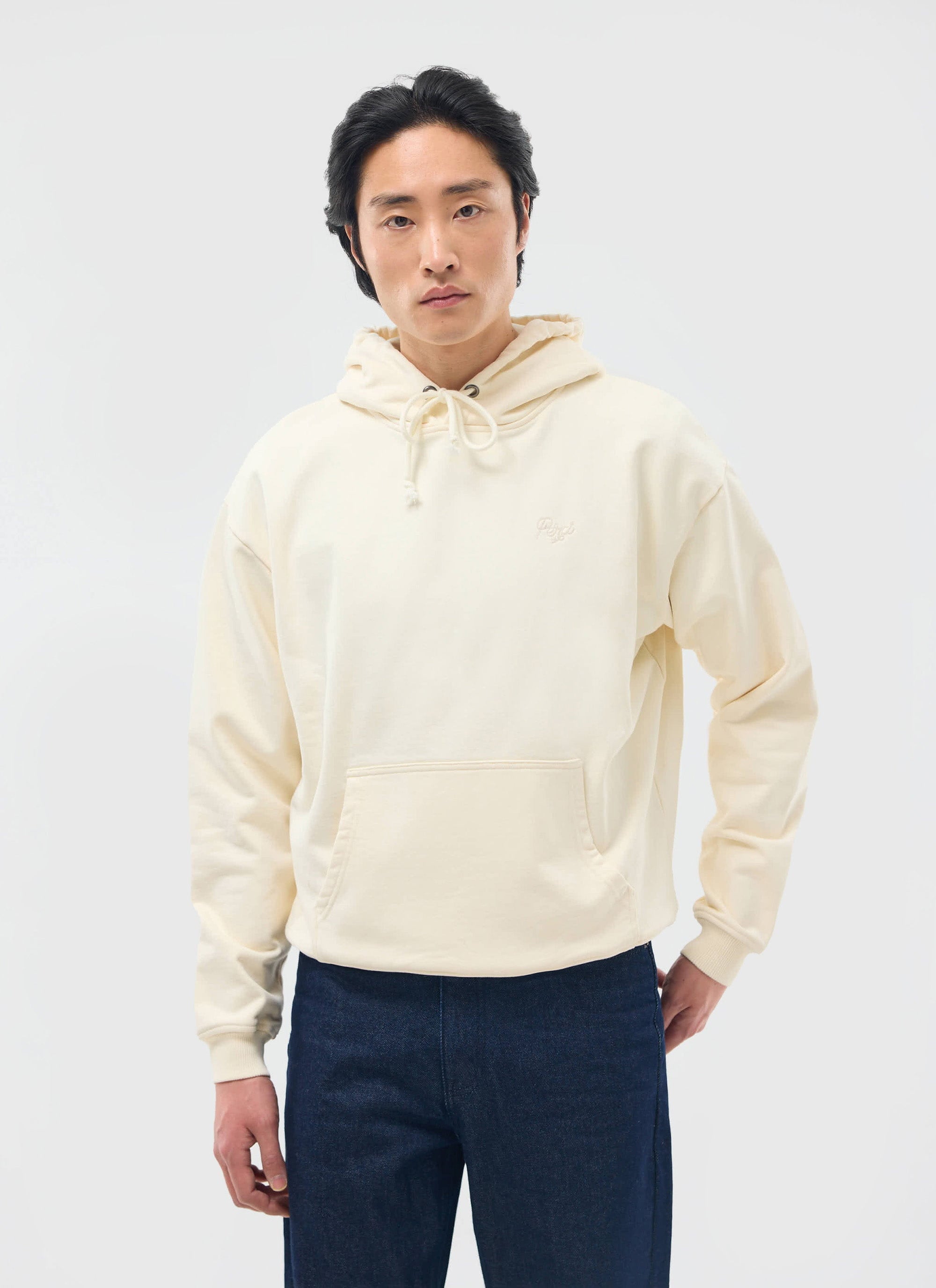 A man models the Percival Perci Chainstitch Hoodie in ecru, showcasing its relaxed fit with dark denim jeans.