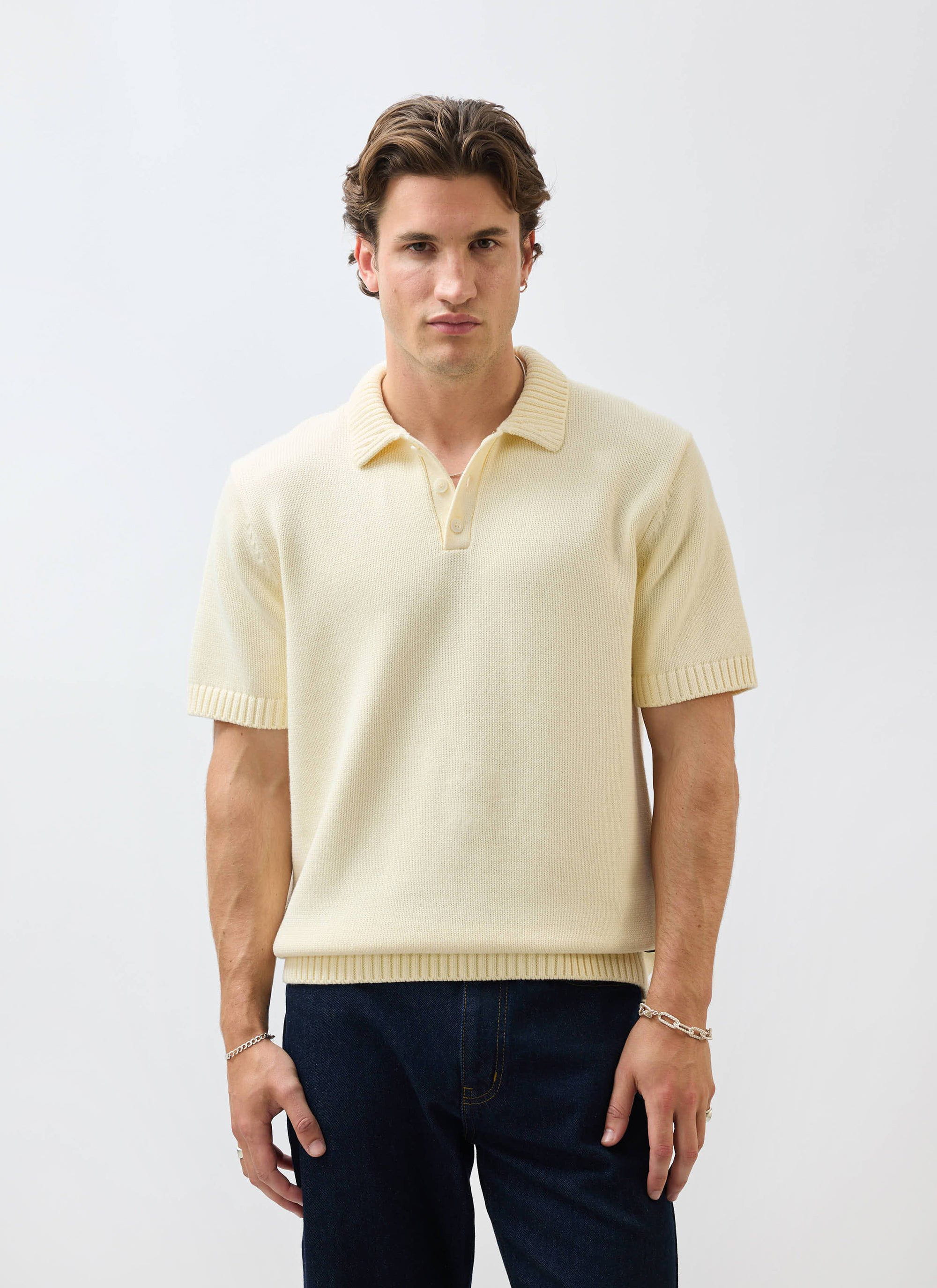 A man models the Percival Verso Short Sleeve Knitted Shirt in ecru, styled with dark jeans against a white background.