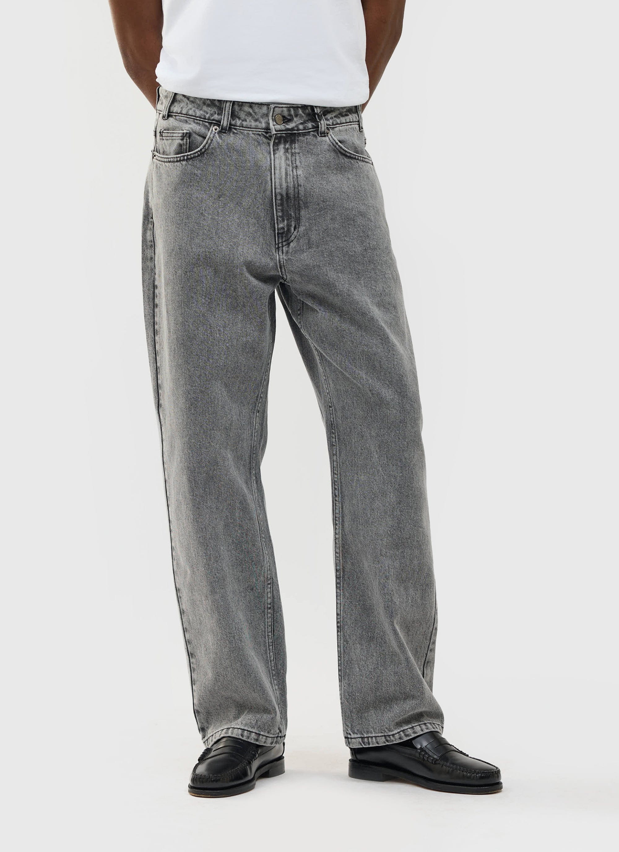 A model wears the Percival Wide Leg Denim Jeans in a warm grey wash, styled with a white tee and black loafers.