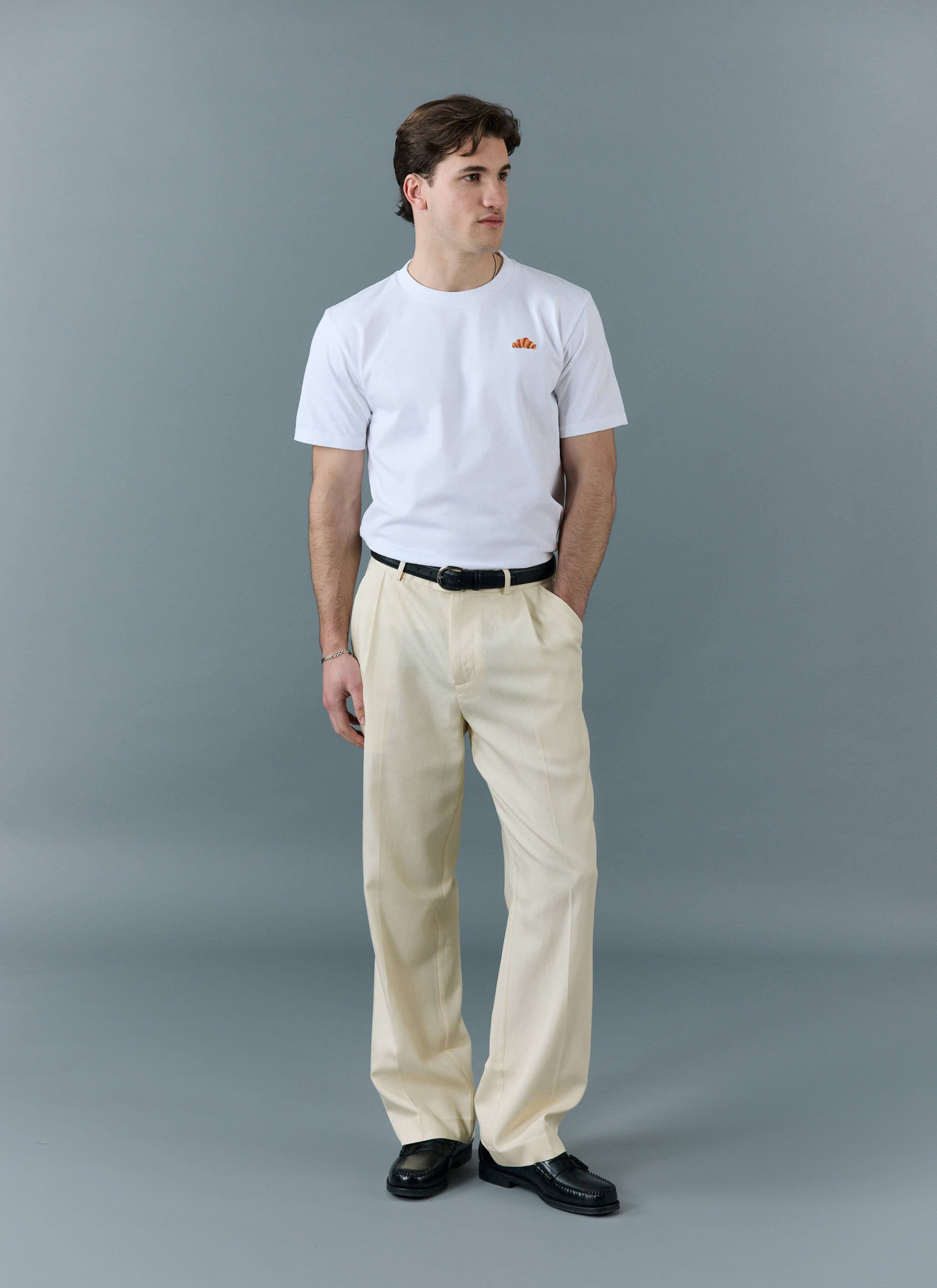 A model wears the Percival white Croissant T-Shirt, tucked into cream trousers with a black belt, against a grey background.