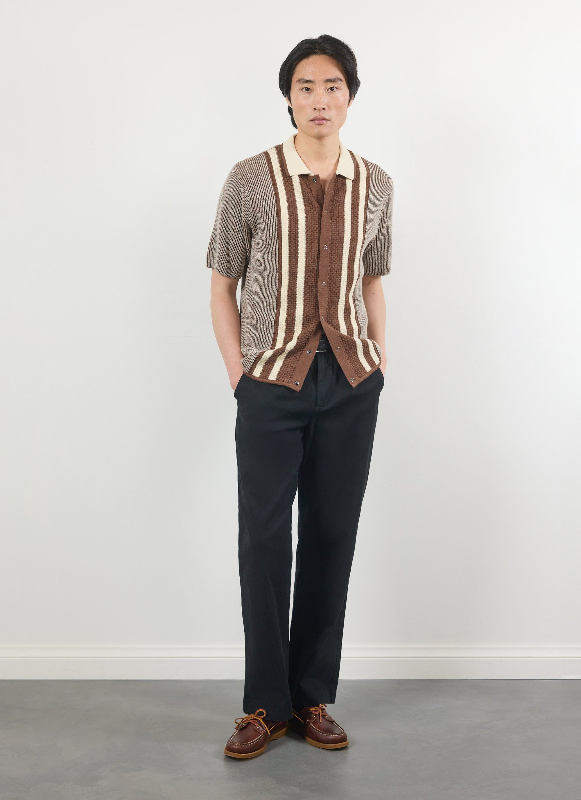 A man models the Percival Teramo Knitted Polo Shirt in ecru, a vertically striped knit styled with black trousers.