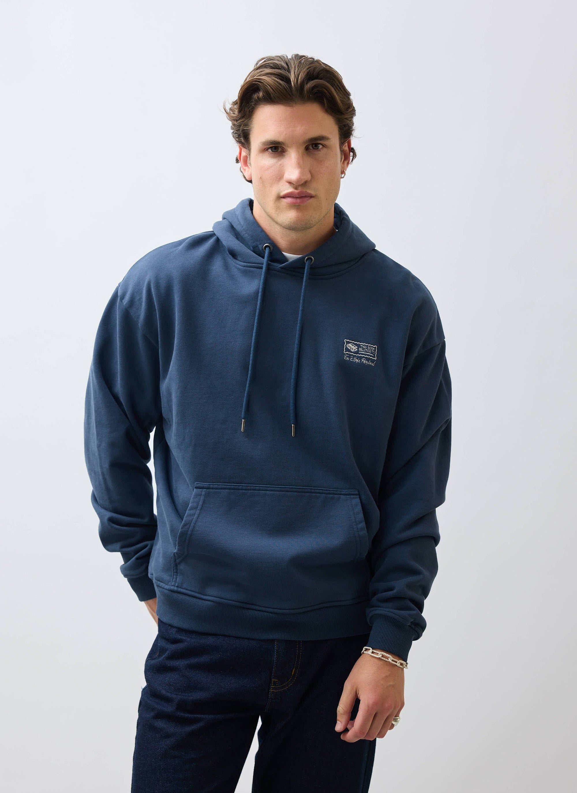 A male model wears the Percival 'This Book Belongs To' hoodie in navy, showing the intricate chest embroidery.